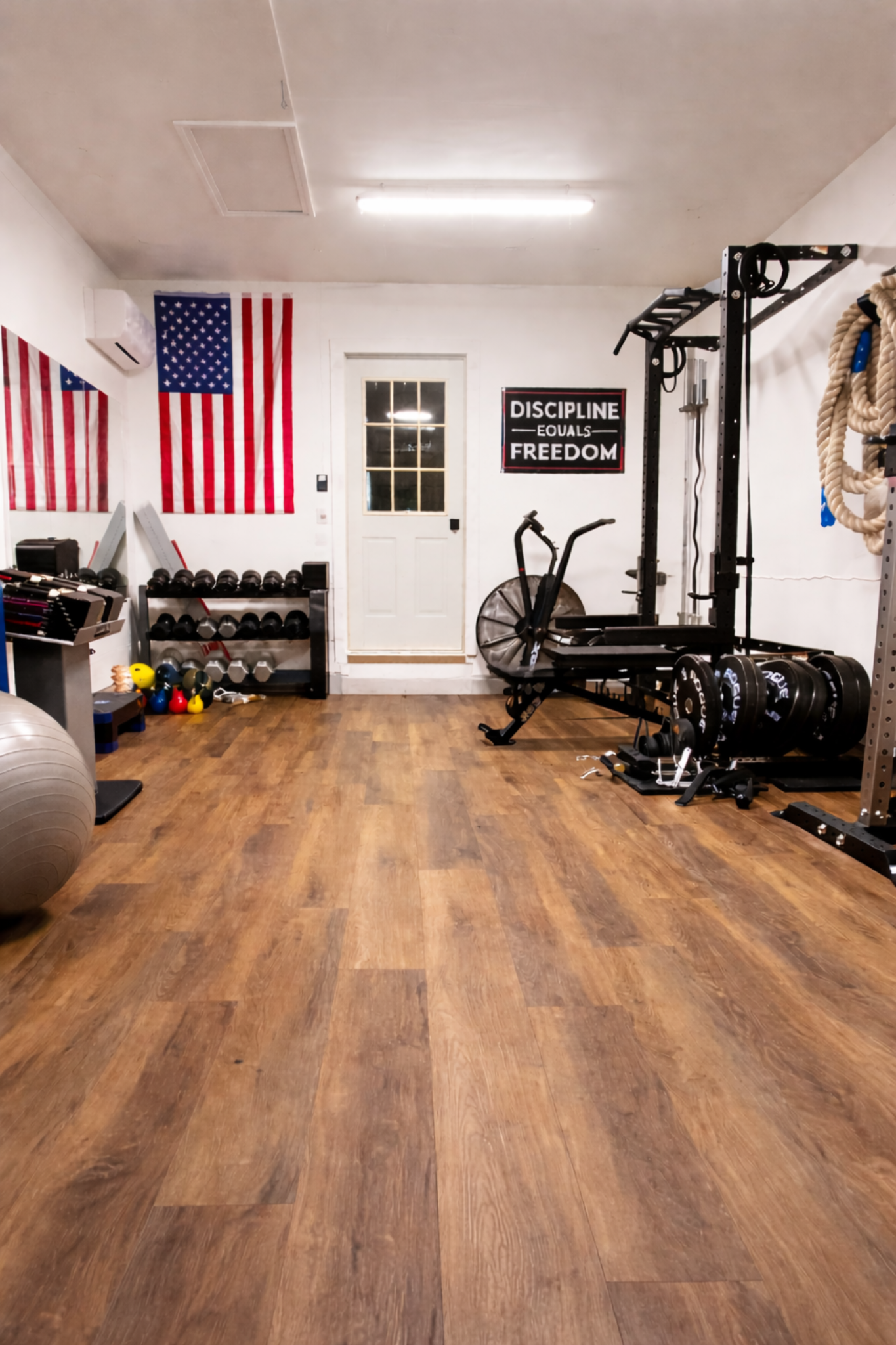 A home gym with American flags on the wall, dumbbells, kettlebells, a fan bike, weight rack, gymnastic rings, and workout equipment on a wooden floor.