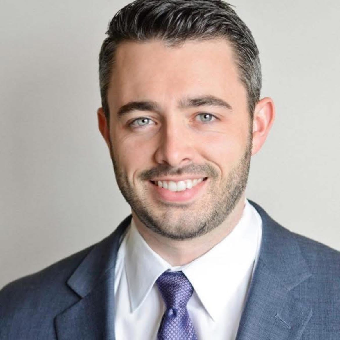 A smiling man dressed in a dark suit, white shirt, and purple tie, with short dark hair and light skin, posing against a plain background.