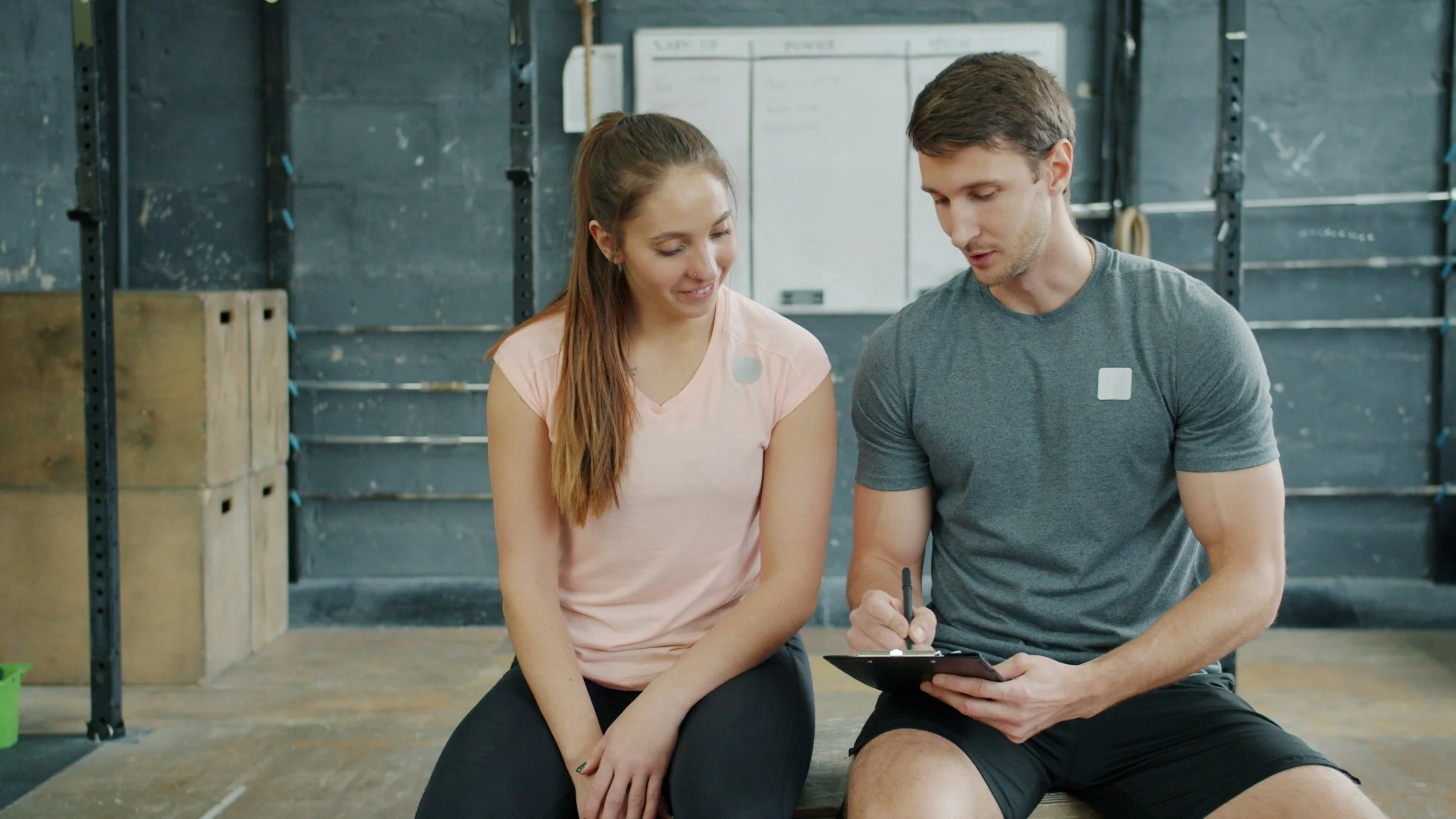 A man and a woman sitting together in a gym or workout space, looking at a clipboard and talking. The woman has long brown hair, is wearing a light pink t-shirt, and has a nose piercing. The man has short brown hair, is wearing a gray t-shirt, and is holding a pen. The background includes gym equipment and a whiteboard.