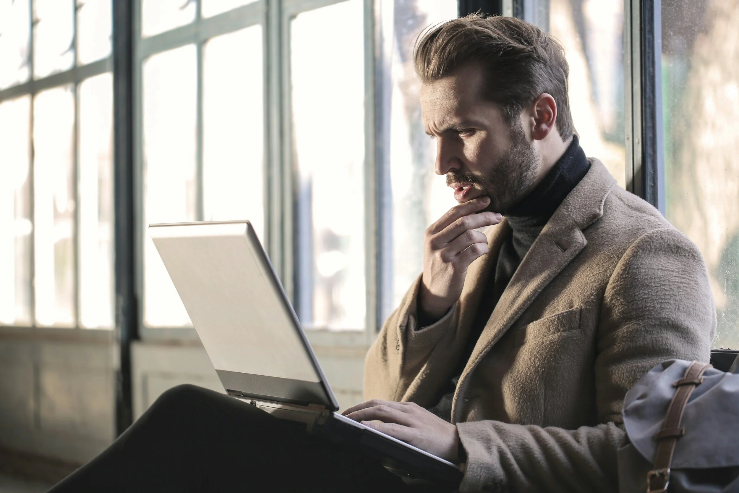 A young man with brown hair and a beard, wearing a brown blazer and black turtleneck, sitting by a window while looking at a laptop, with one hand on his chin, inside a well-lit space with large windows.