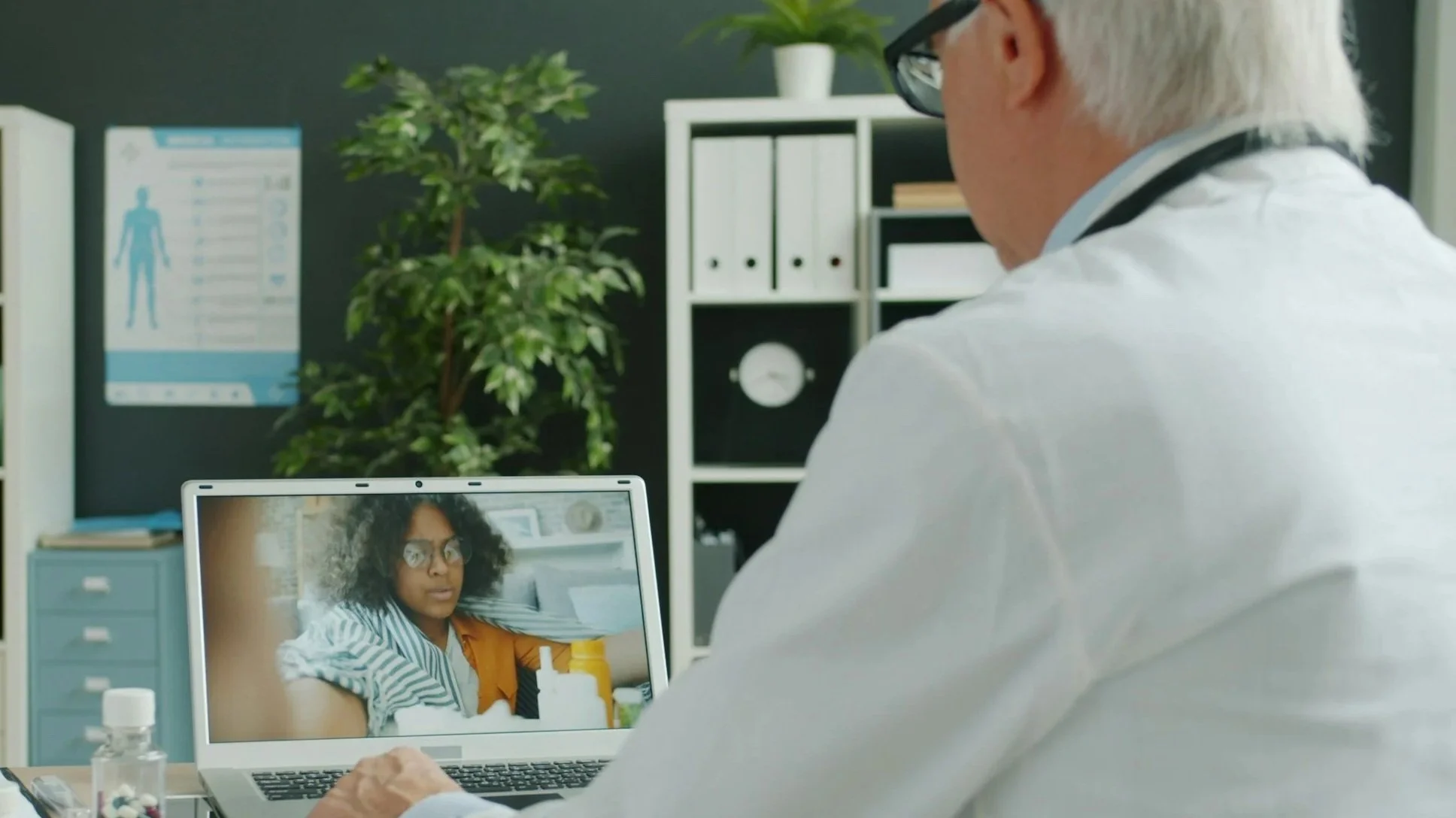 Doctor in a white coat and glasses consulting a female patient via video call on a laptop in an office with shelves, plants, and a wall poster.