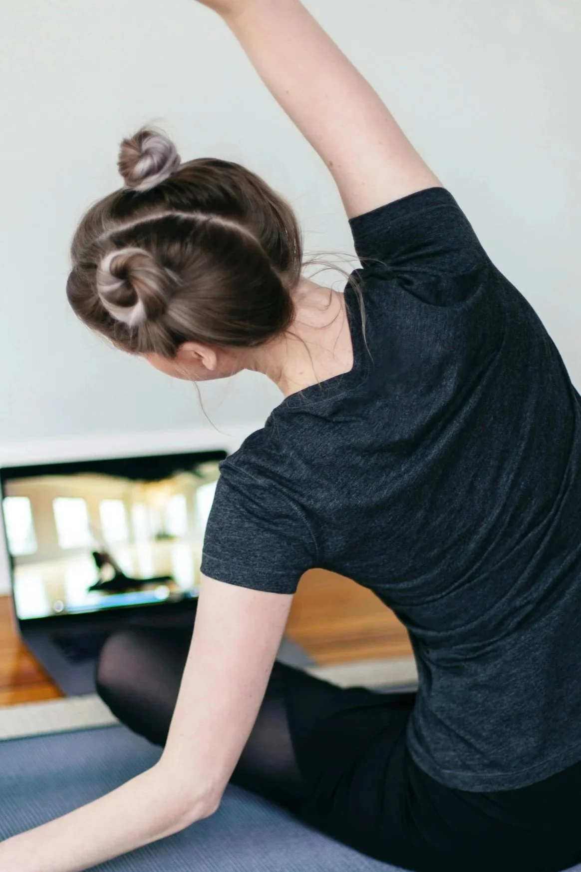 A woman practicing yoga or stretching indoors, with a laptop in the background showing a person sitting on a mat.