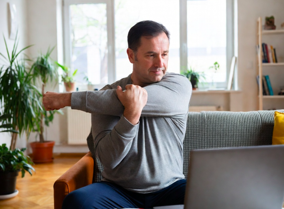 A man stretching his arm outside of a gray long sleeve shirt while sitting on a couch in a living room with plants and a bookshelf in the background.
