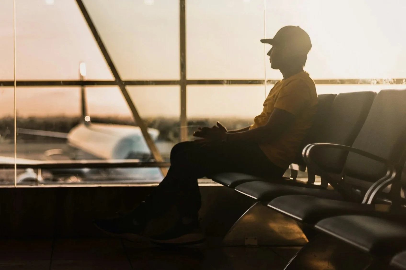 Silhouetted person sitting on airport bench near window, with a view of an airplane and sunset outside.