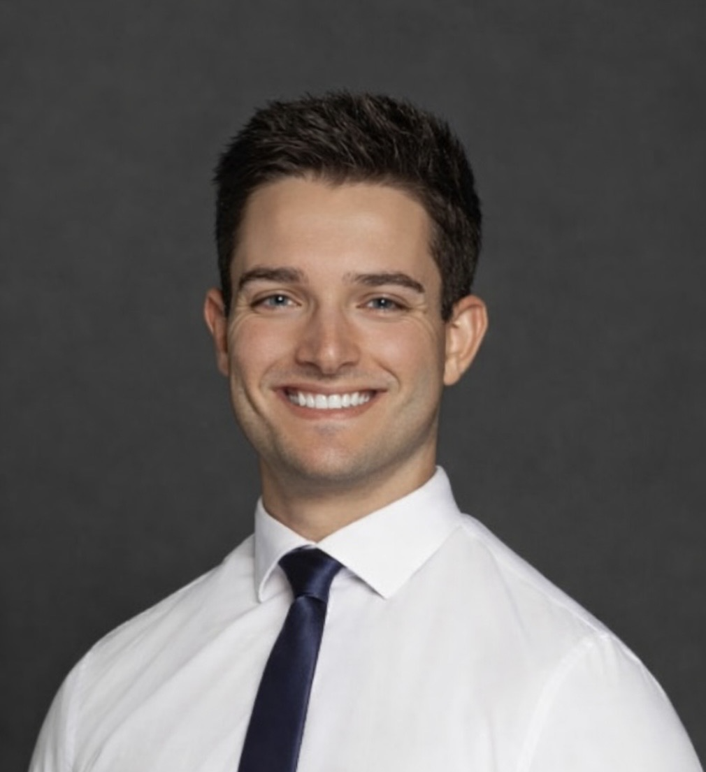 A young man with dark hair, wearing a white shirt and a dark tie, smiling against a dark gray background.