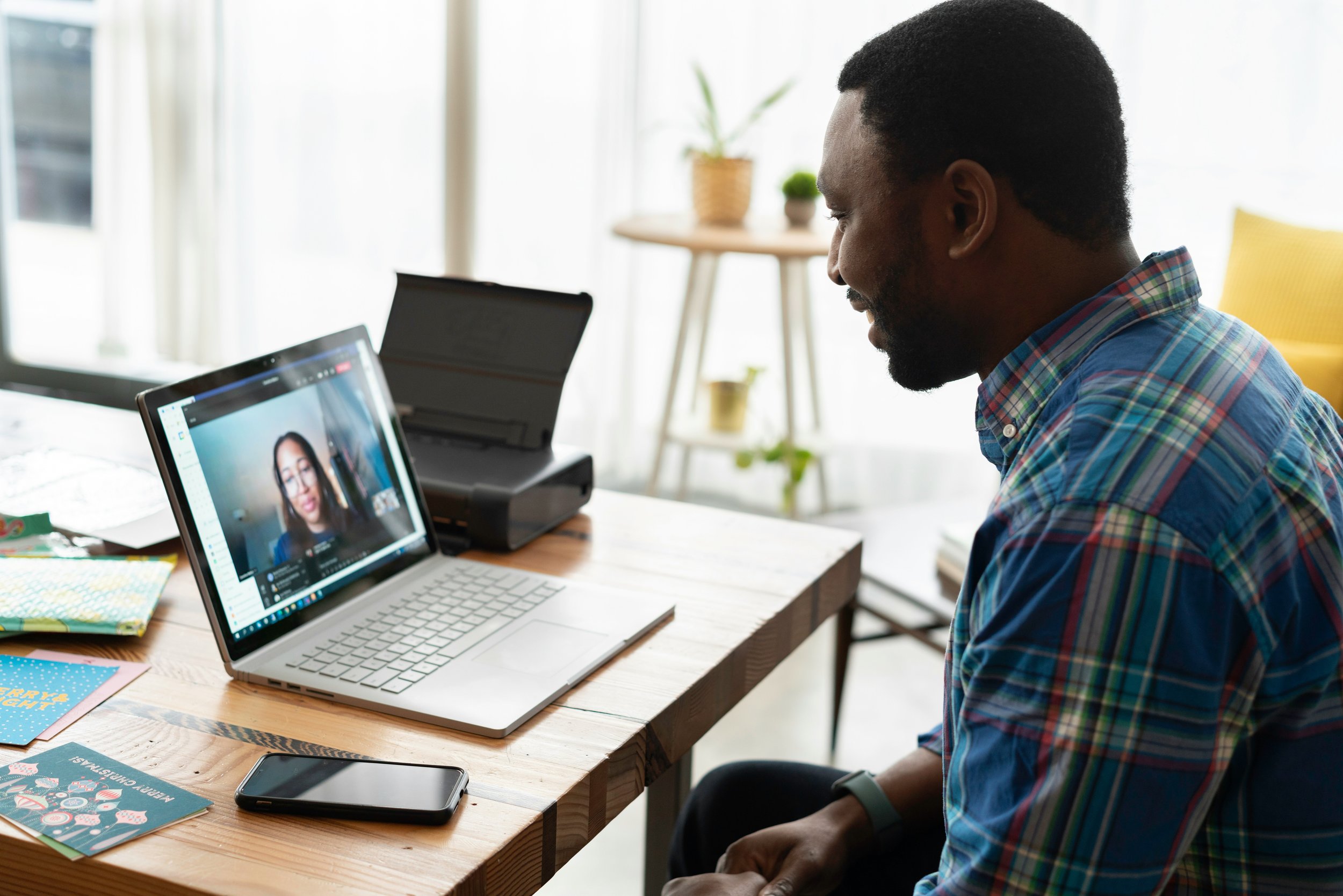 A man in a plaid shirt participating in a video call with a woman on a laptop, sitting at a wooden desk with colorful greeting cards, a smartphone, and office supplies, in a bright room with large windows.