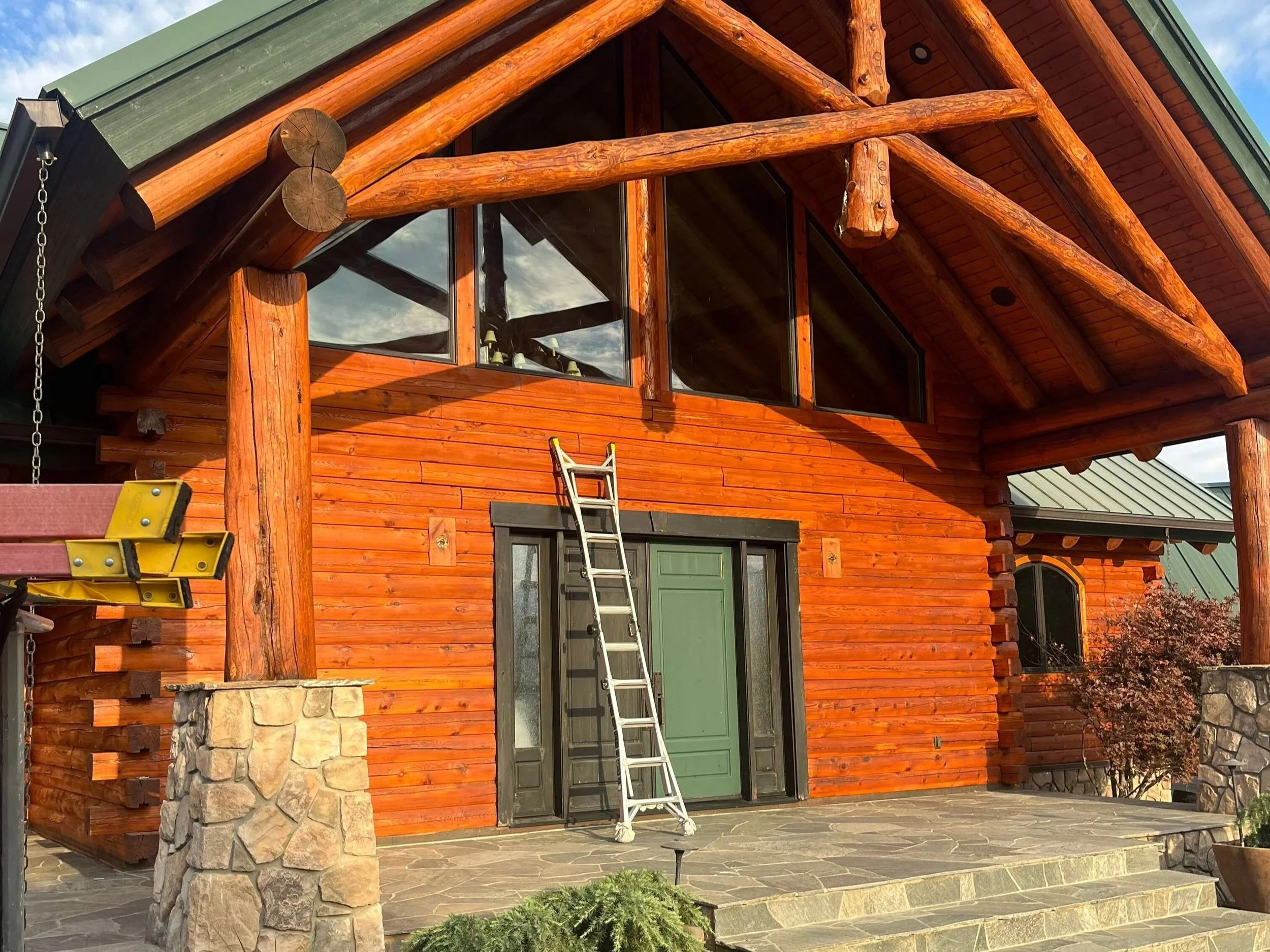 Front view of a wooden log cabin with large green door, stone porch, and large windows under a covered entrance. A ladder is leaning against the door, and there is a tree to the right side.