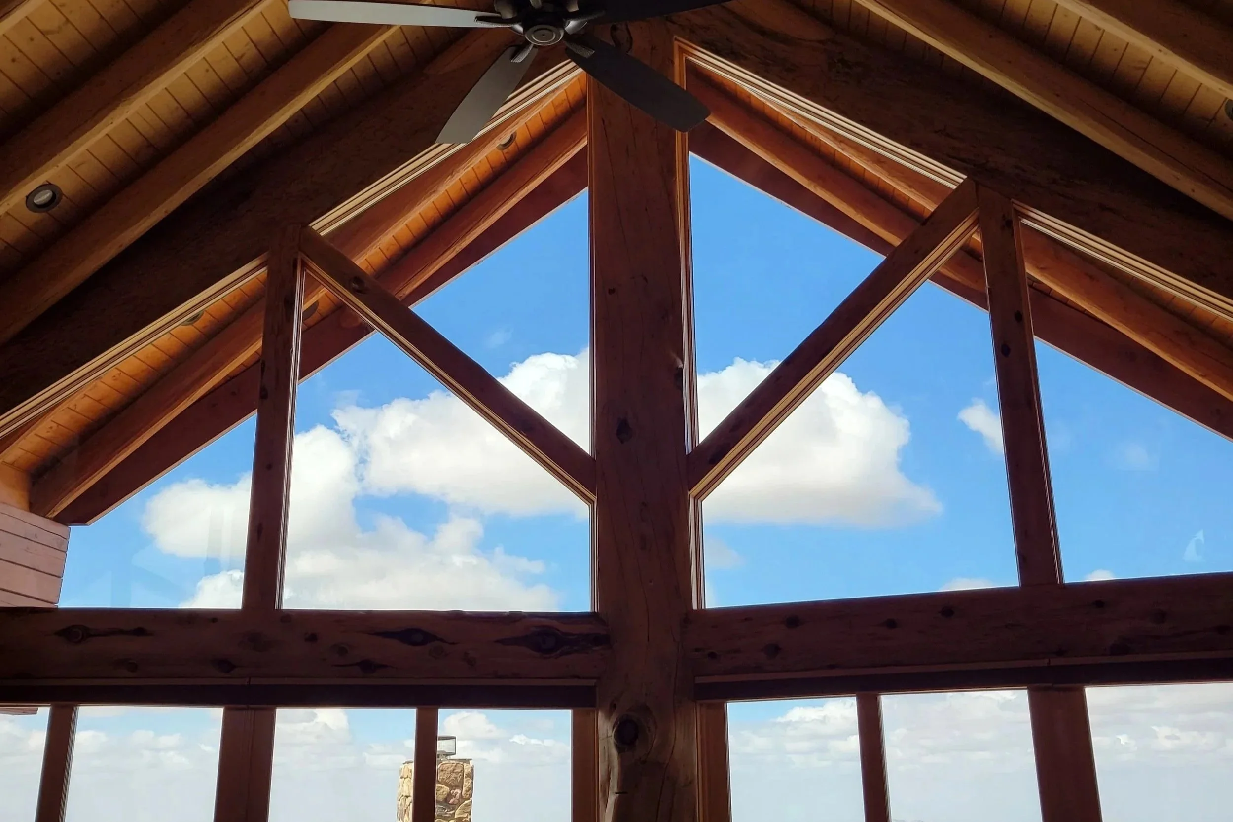 Looking up at the interior of a wooden attic space with large windows showcasing a bright blue sky with scattered clouds.