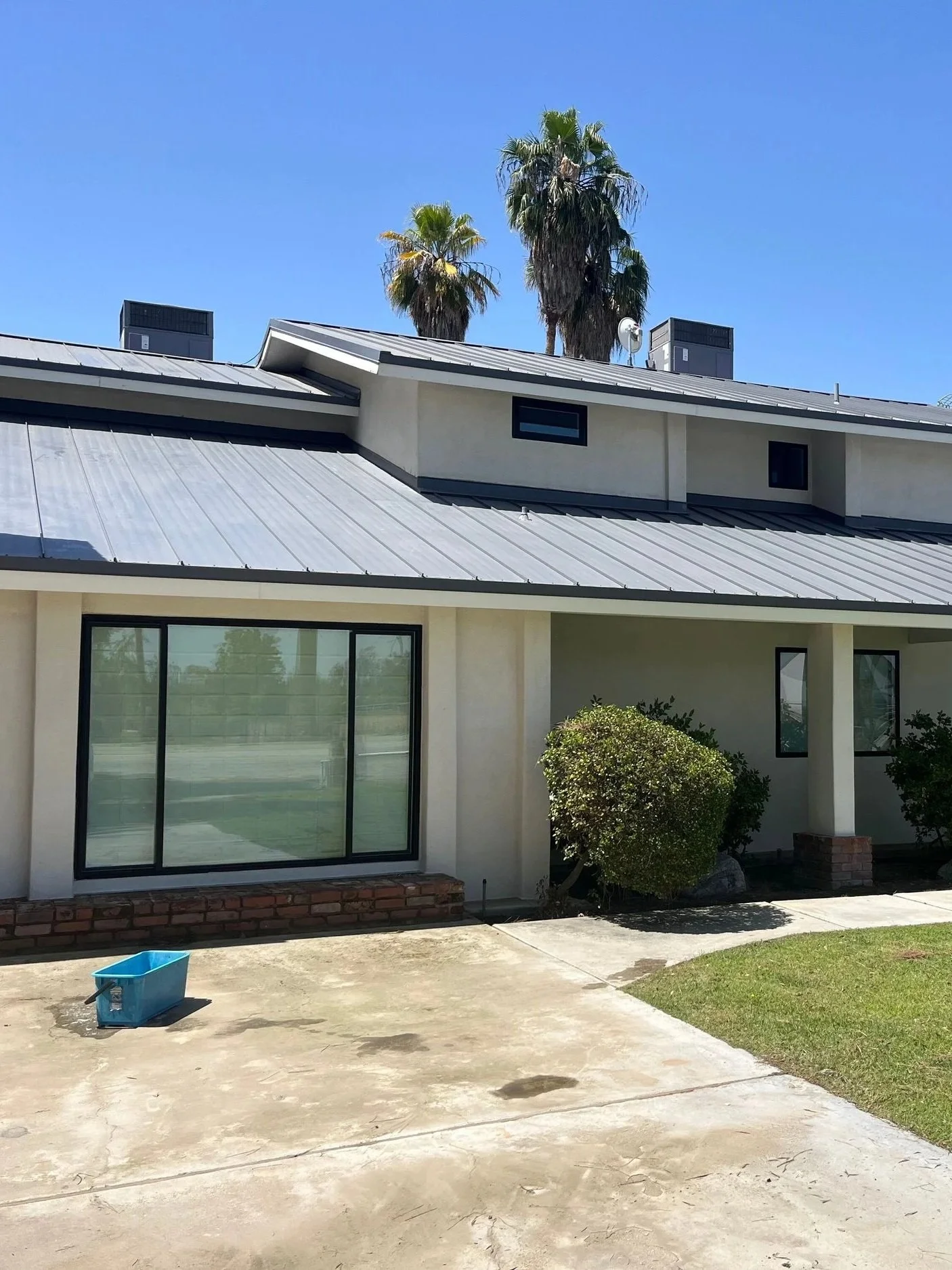 View of a modern house with beige exterior walls, large sliding glass window, metal roof, small bushes, and a concrete driveway under a clear blue sky with tall palm trees in the background.