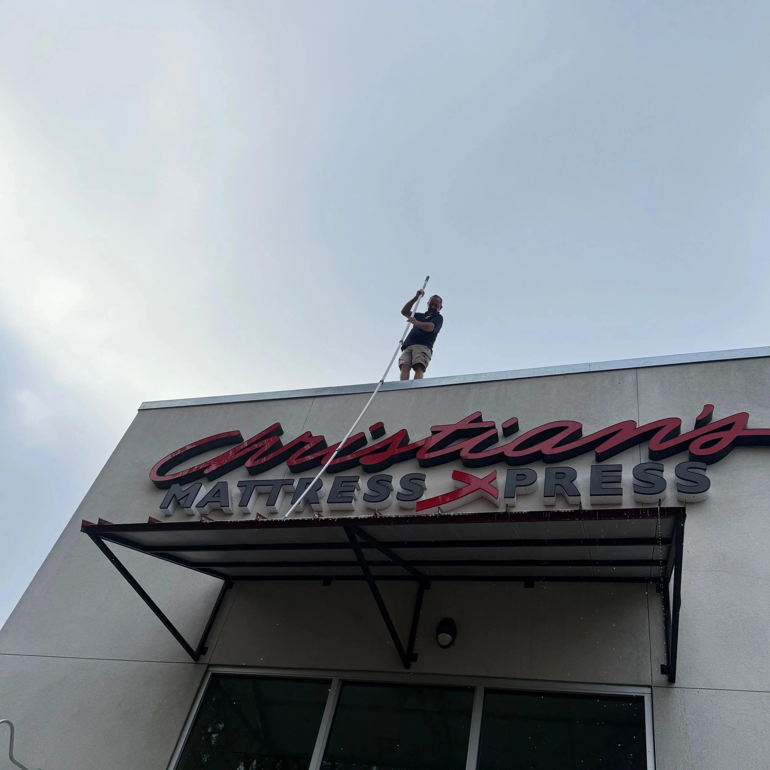 A person cleaning or maintaining the sign on top of a Mattress Xpress store using a long pole and a harness for safety.