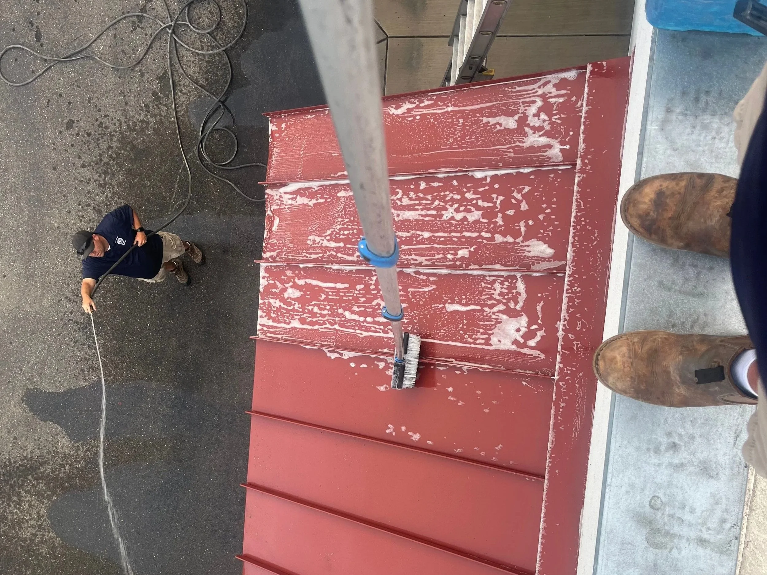Person cleaning a metal red roof using a roller brush, viewed from above with another person spray washing the ground below.