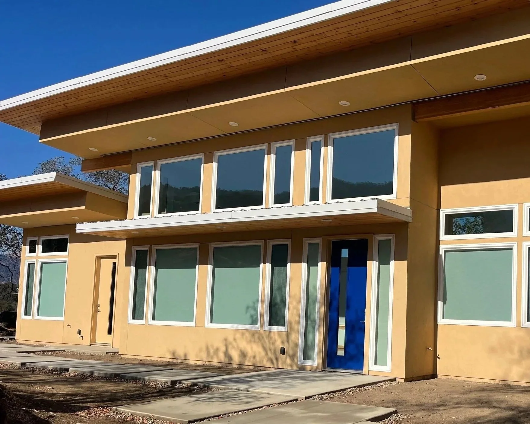 Modern two-story house under construction with large windows, tan exterior walls, and a flat roof, set against a clear blue sky.