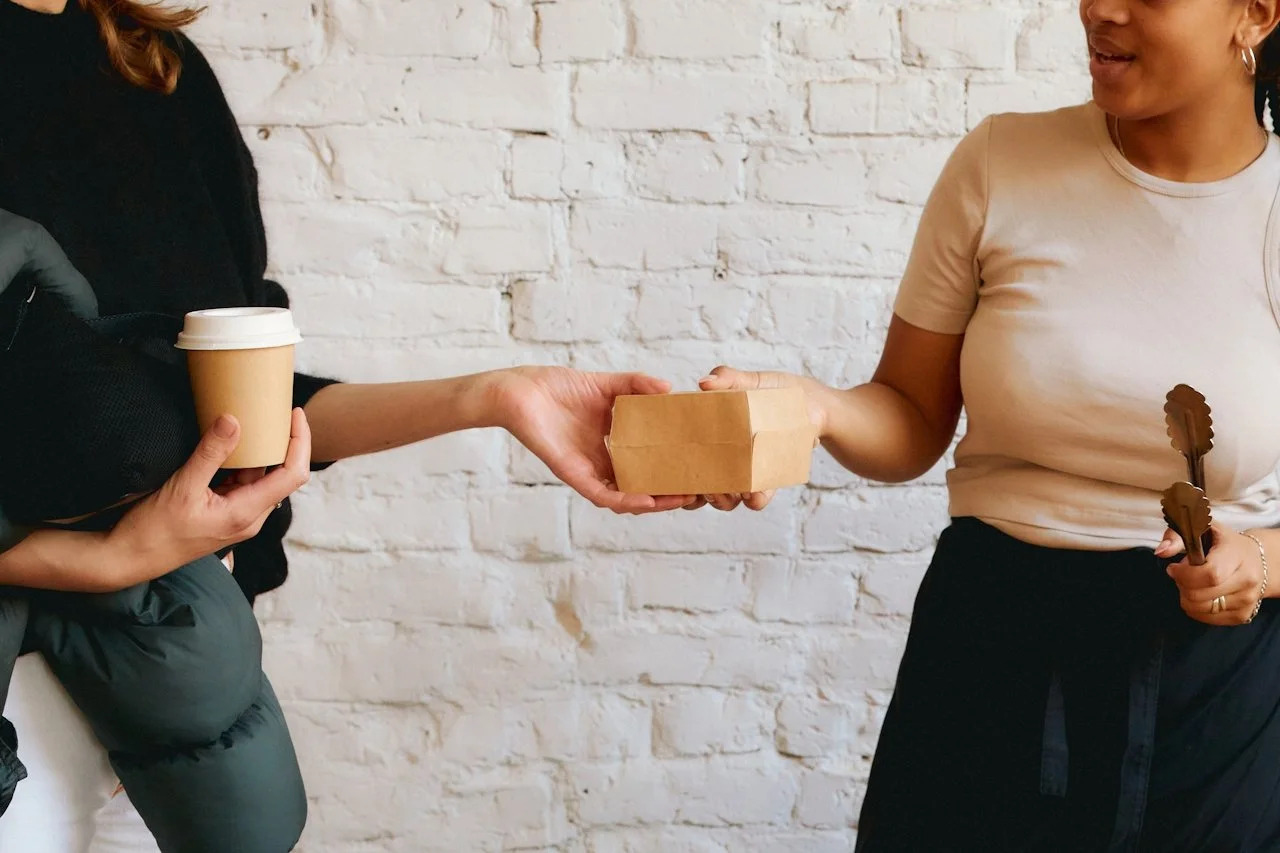 Person on left handing a takeout box to person on right, who is holding tongs, while holding a coffee cup.
