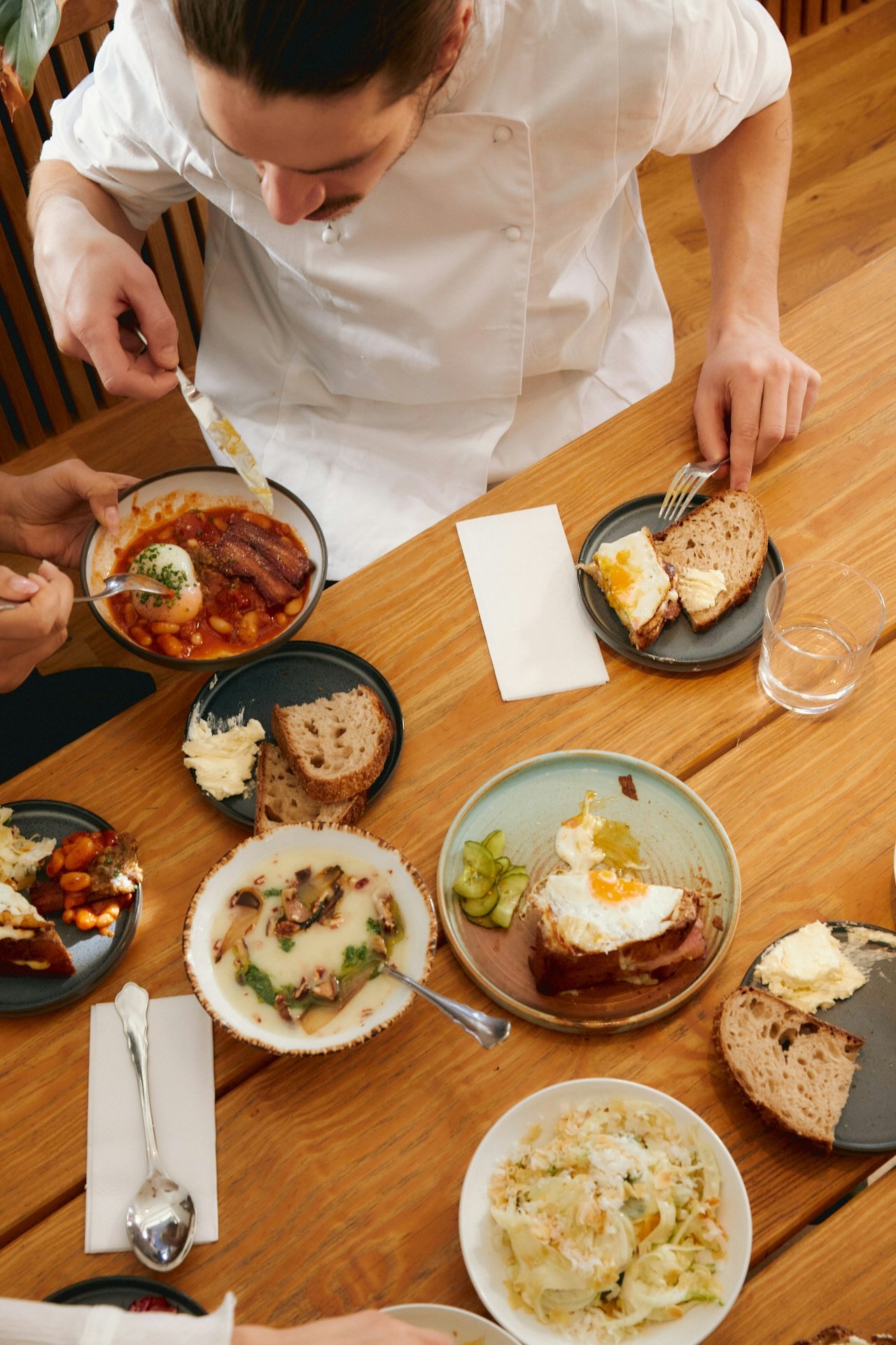 A person in a white chef's coat leaning over a wooden table set with various dishes including bread, eggs, stews, salads, and spreads, with utensils and napkins