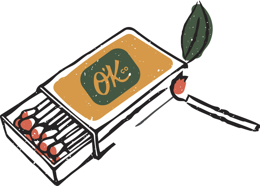 A grill with a cheese burger labeled "OK Co" on top and a spatula with a leaf, with some sausages cooking on the grill.