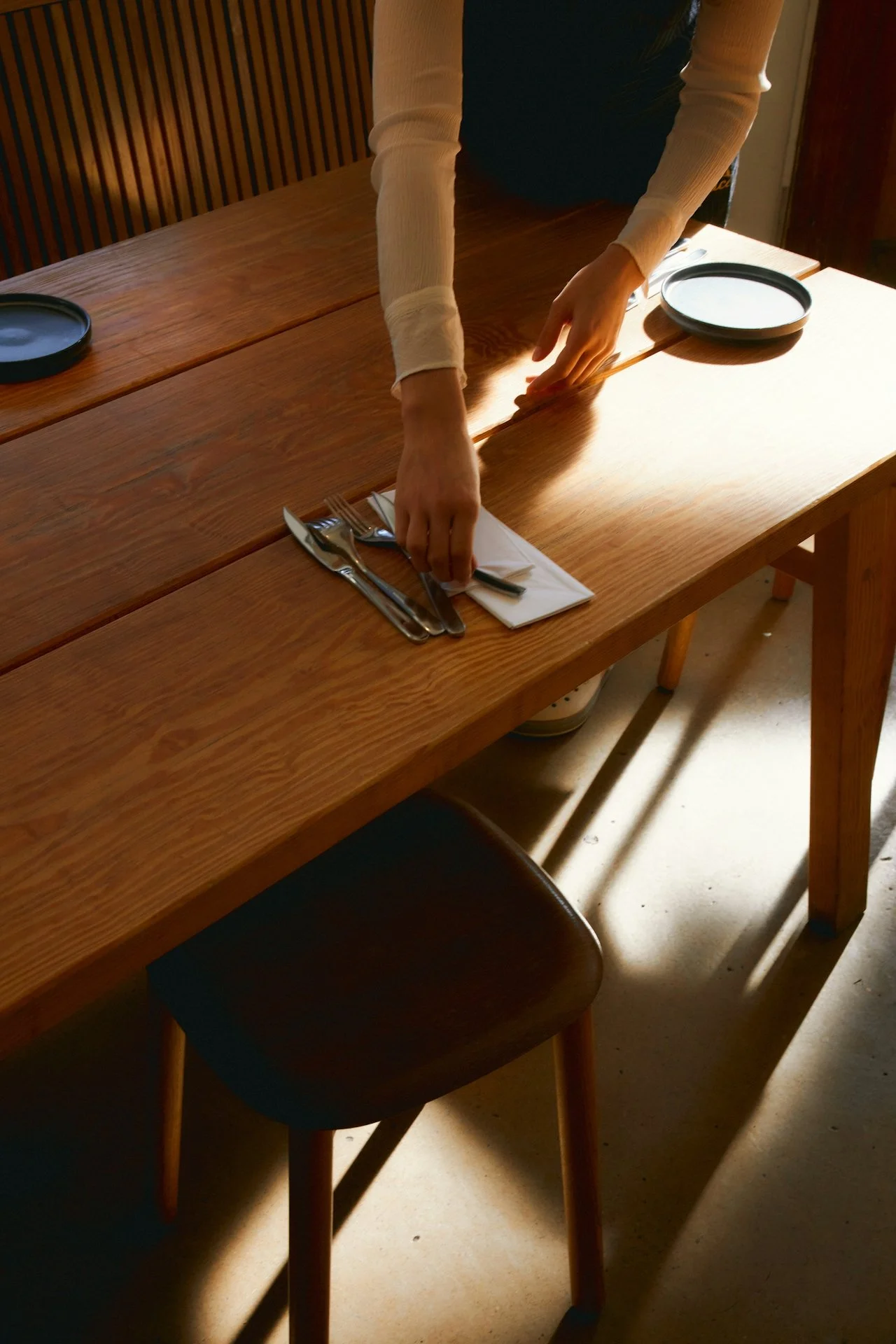 Person setting a table in a dining area, with a wooden table, cloth napkin, fork, knife, and plate, illuminated by natural light.