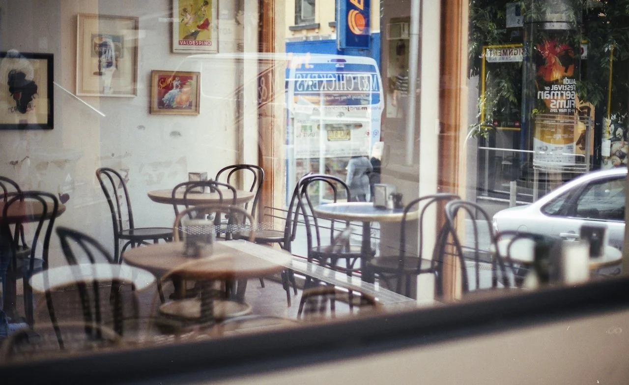 Empty cafe with round tables and chairs inside, seen through a glass window with reflections of street, cars, and posters outside.