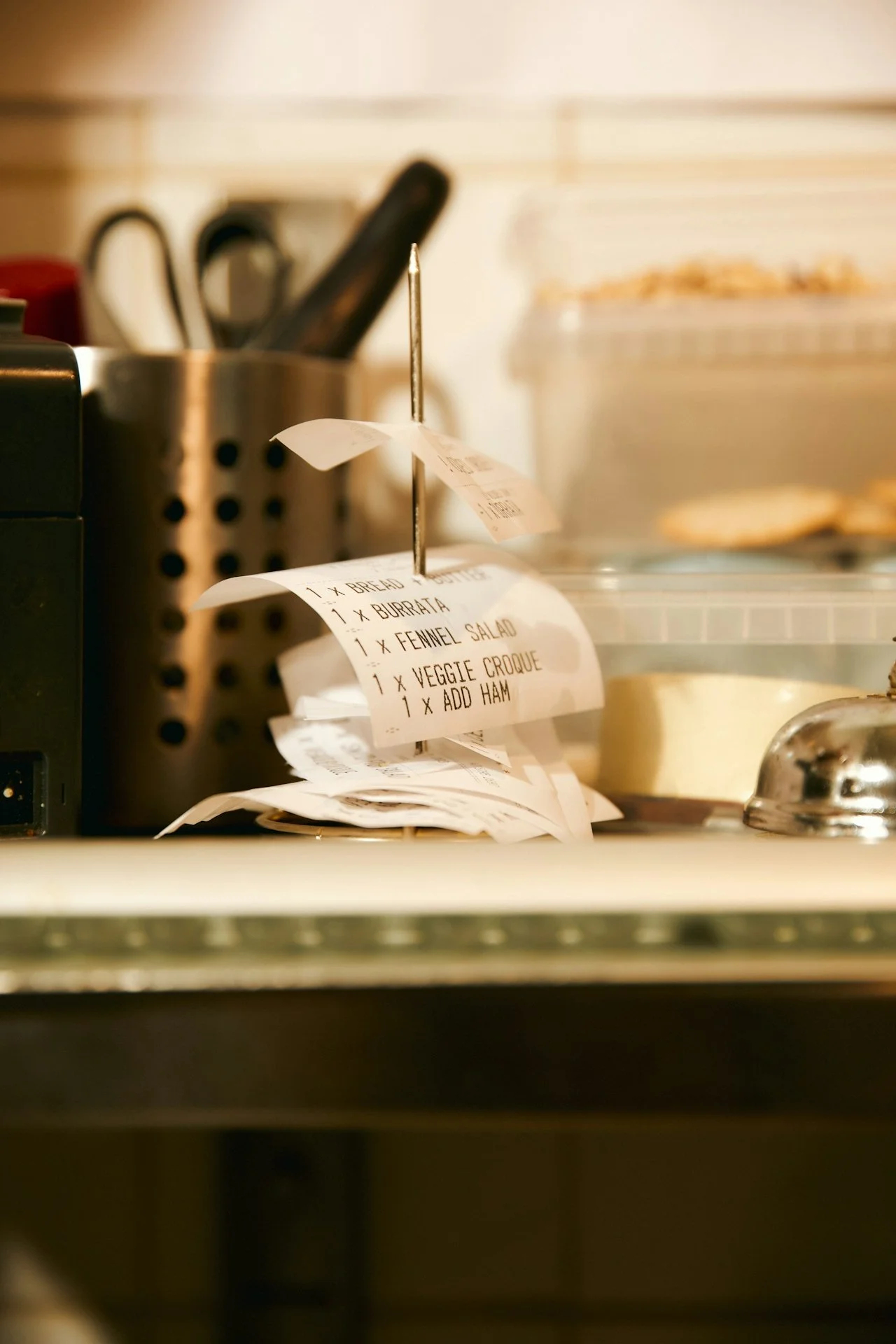 A close-up of a deli ticket with order details hanging from a metal stand, with a blurred background of kitchen utensils and food containers.