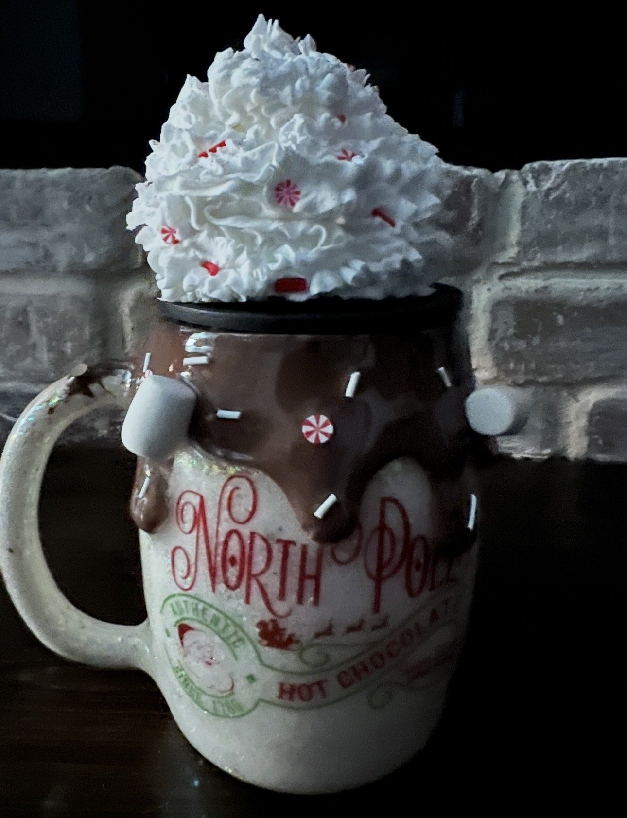 A North Pole-themed ceramic mug filled with hot chocolate, topped with whipped cream and red and white holiday sprinkles, placed against a brick wall.