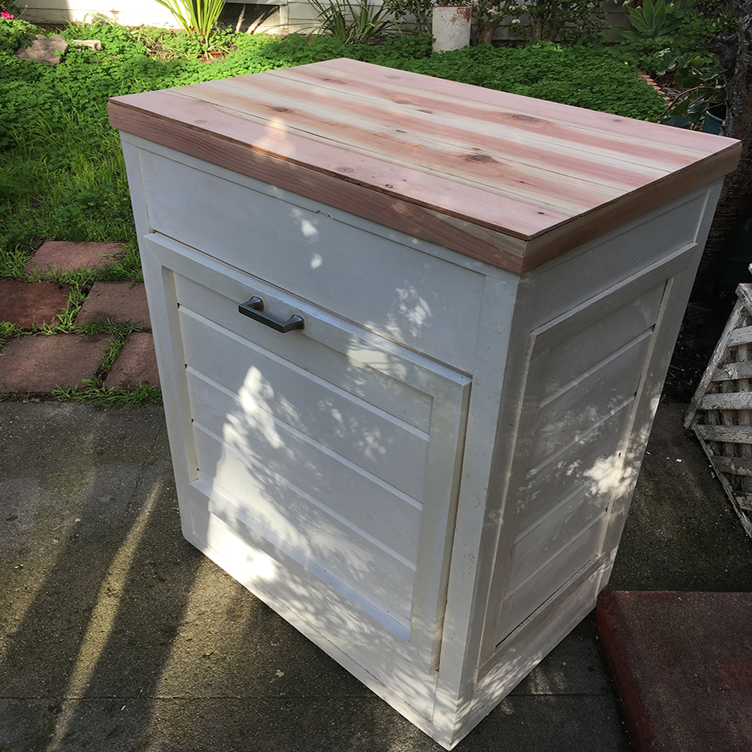 A small white wooden cabinet with a drawer and a door, featuring a black handle, placed outdoors on a concrete surface with plants and greenery in the background. The top of the cabinet is a light reddish-brown wood.