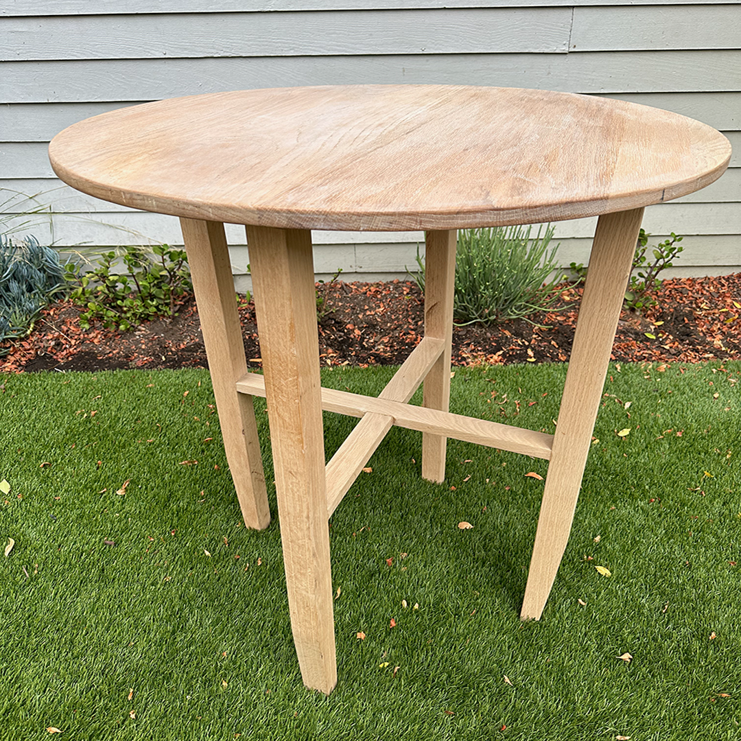 A wooden outdoor table with a round top situated on a grassy lawn with flower bed and house siding in the background.