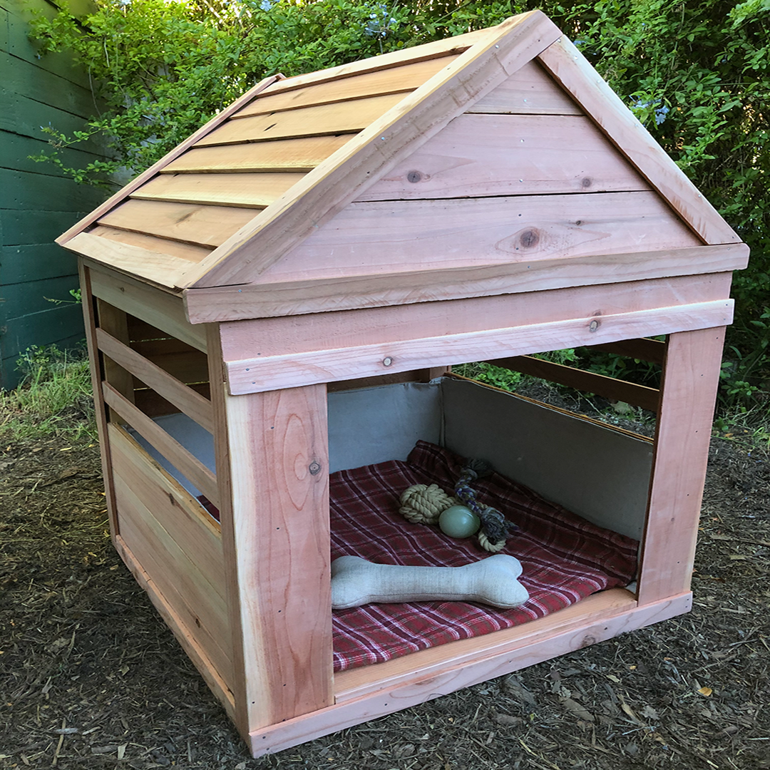 Wooden dog house with a sloped roof and an open front, inside are dog toys and a blanket, set outdoors on soil with greenery in the background.