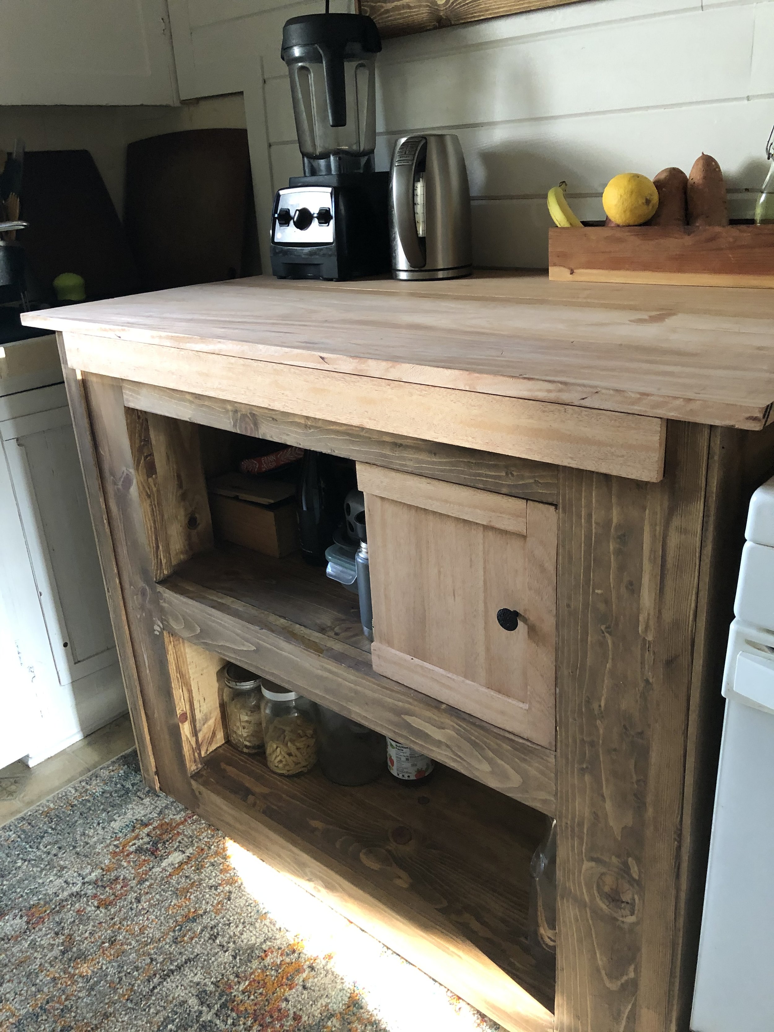 A wooden kitchen cabinet with open shelves, containing jars of pasta and other food items, a blender, a toaster, a banana, a lemon, and sweet potatoes on top, with a textured multicolored rug in front.