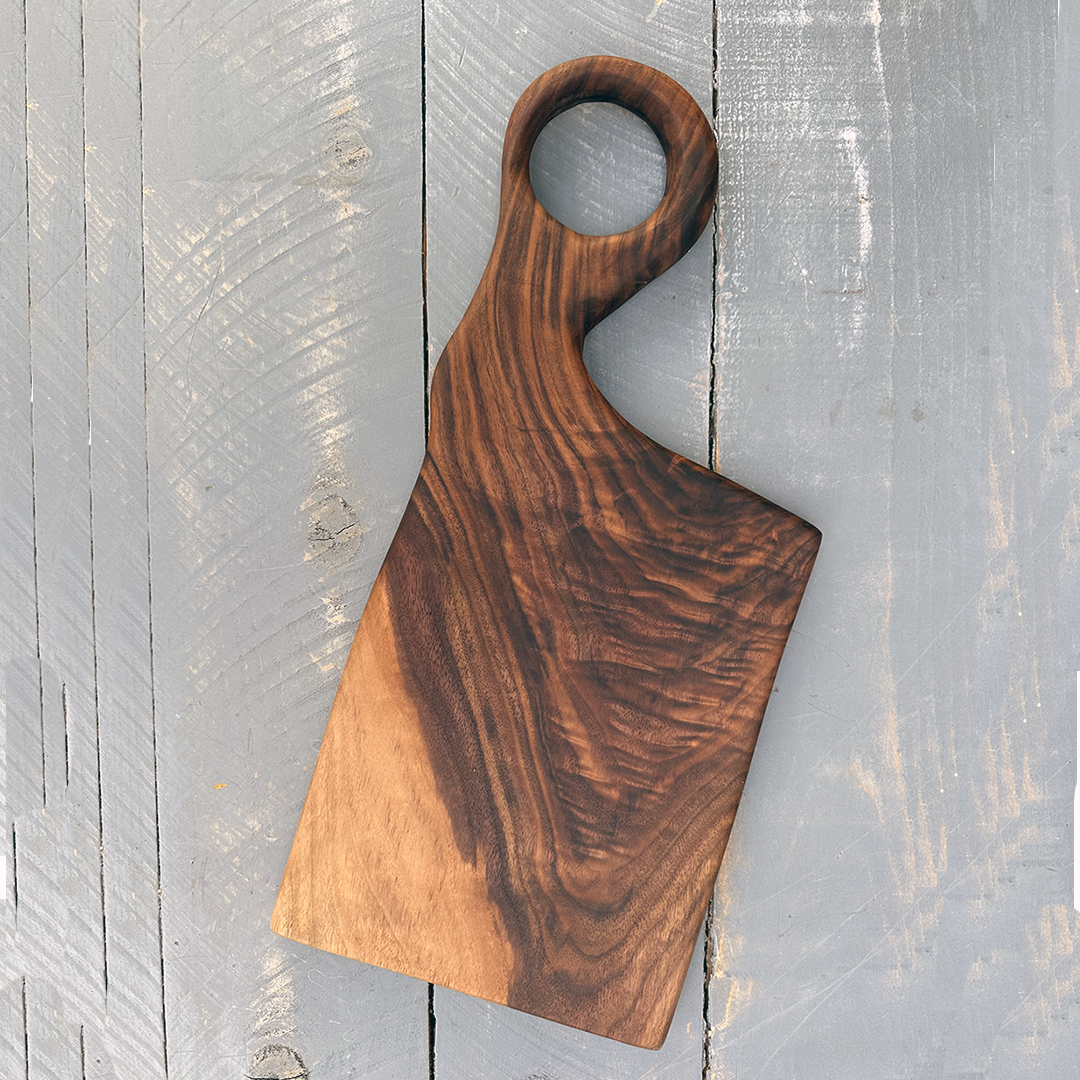 A wooden cutting board with a handle and a circular hole, resting on a weathered gray wooden surface.