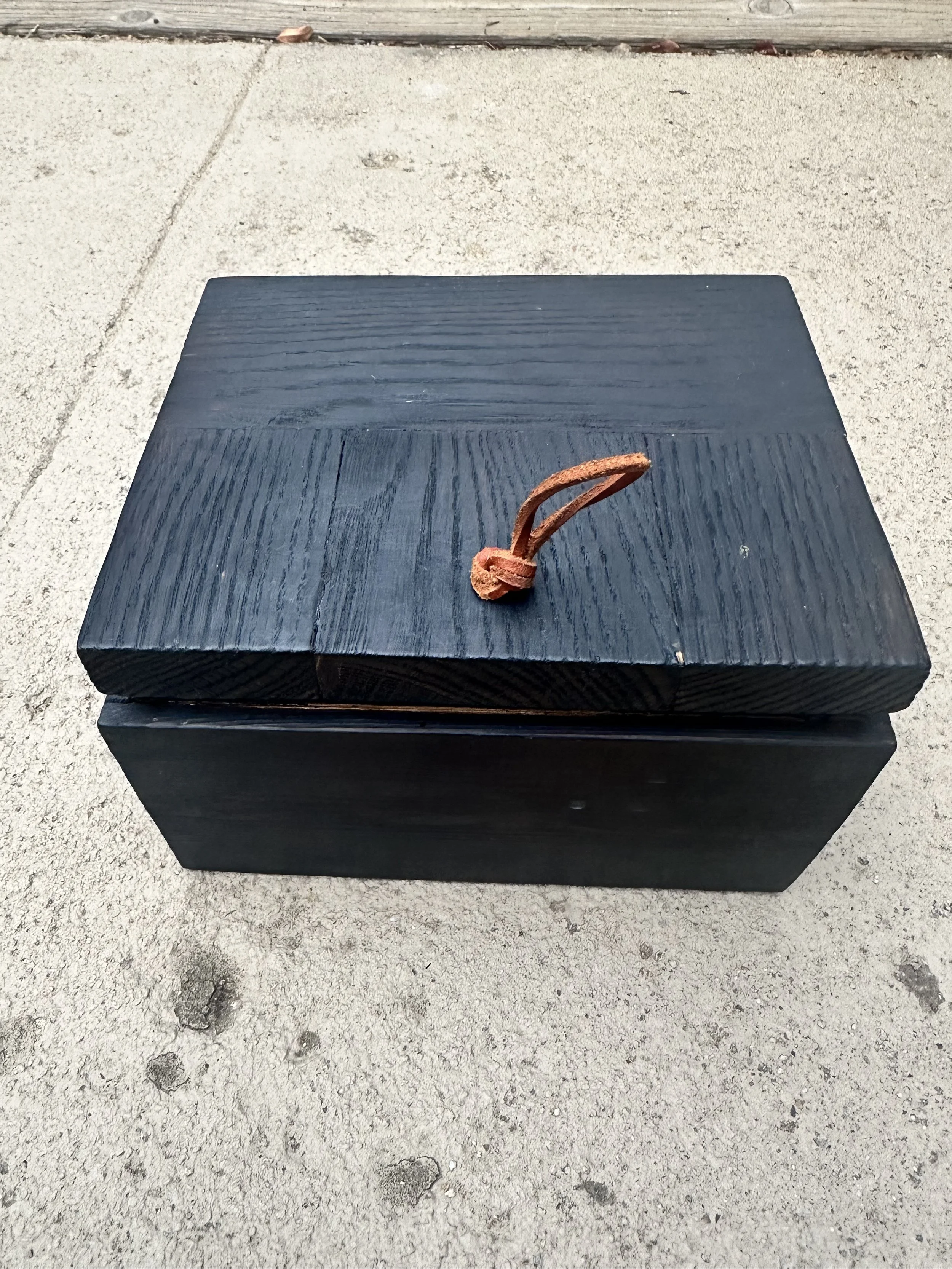 A black wooden bench on concrete pavement with a rusted metal hook on top of it. The background includes a wooden board and some small debris on the sidewalk.