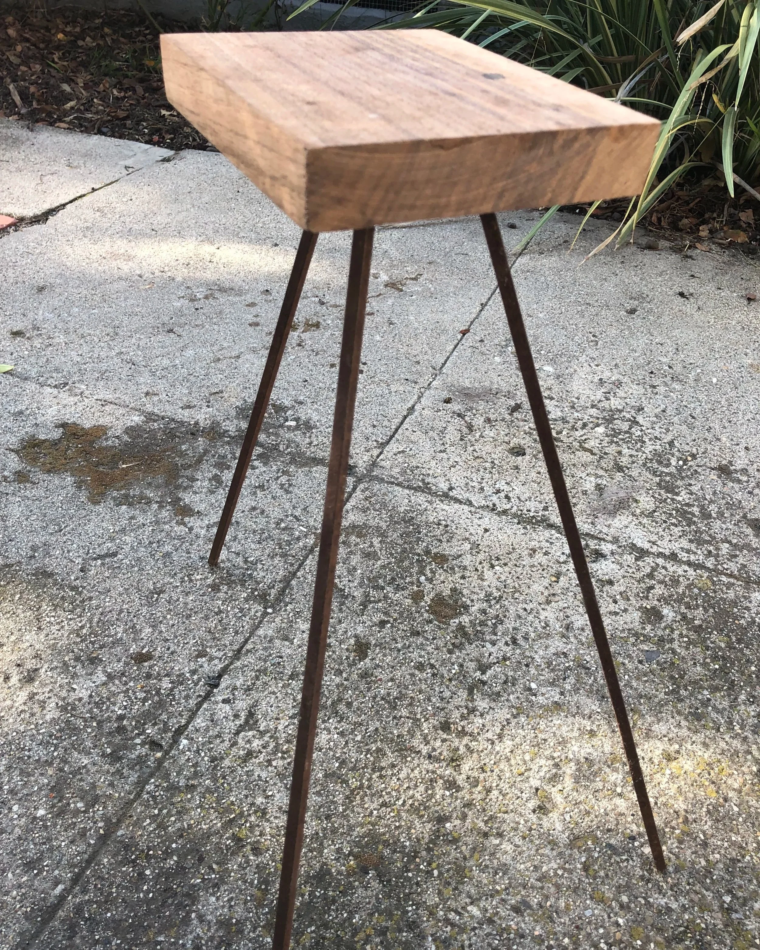 A small wooden table with three metal legs standing on a concrete surface outdoors. Green plants are visible in the background.