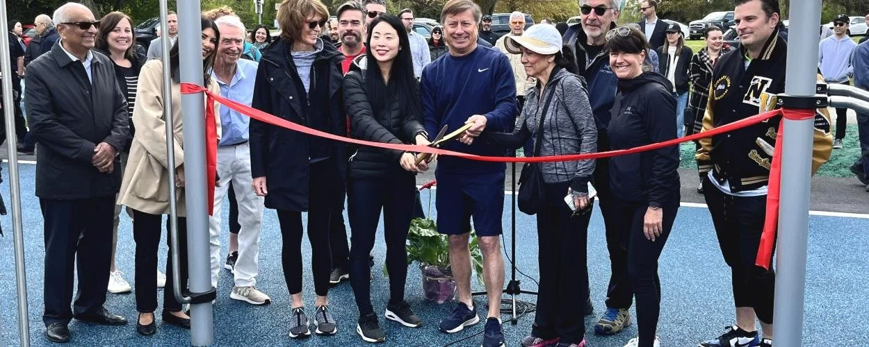 Group of people at an outdoor ribbon-cutting ceremony, standing on a blue surface, with trees and parked cars in the background.