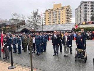 Group of uniformed personnel and officials standing outdoors on a cloudy day, with buildings and a parade float in the background.