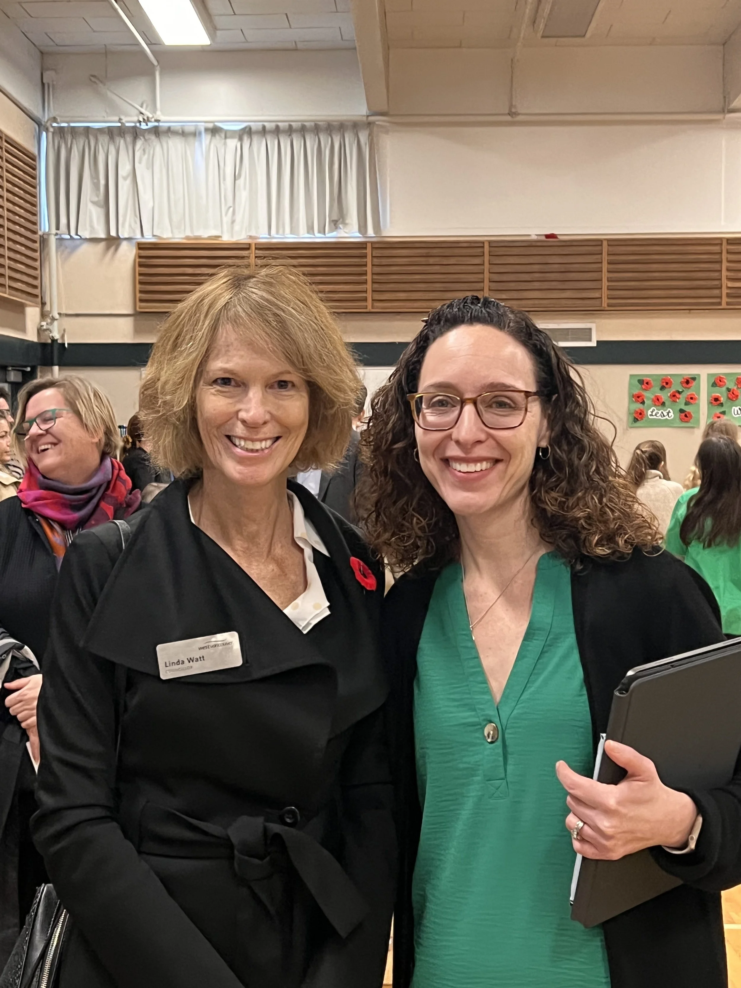 Two women smiling at an indoor event, with other people in the background. The woman on the left has short, light brown hair, and is wearing a black jacket with a name badge. The woman on the right has long, curly dark hair, glasses, and is wearing a green top with a black blazer and holding a tablet or notebook.