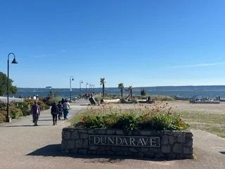 People walking along a pathway by the beach in Dundarave, with a stone planter and a view of the ocean and distant hills under a clear blue sky.
