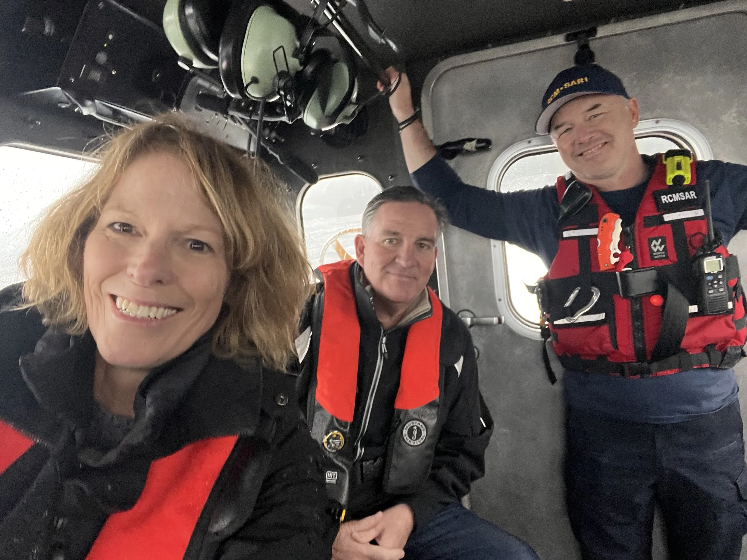 Three smiling people inside a helicopter, with one person wearing a red safety vest and a headset, and another wearing a black cap with yellow writing, all posing for a photo.