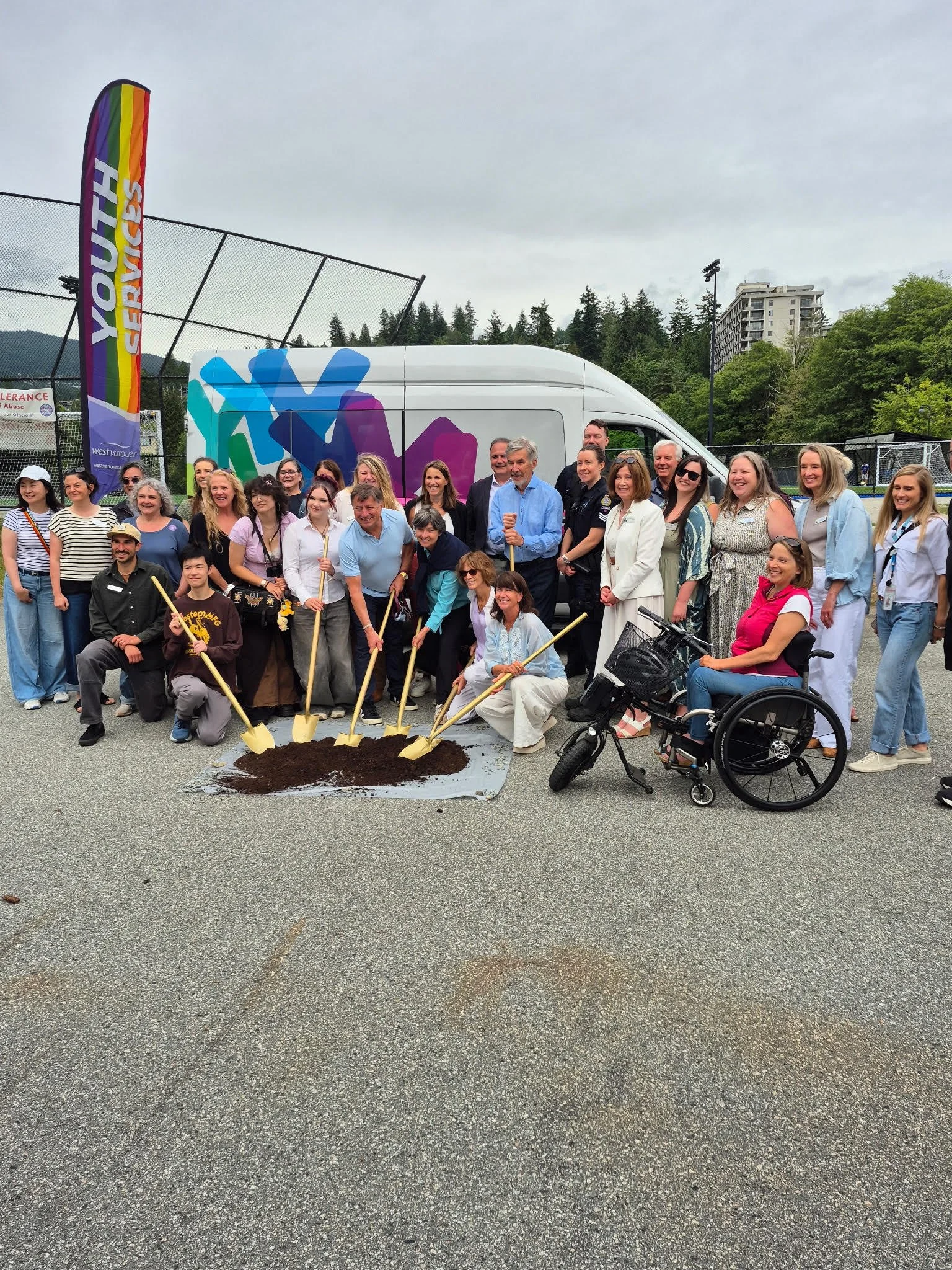 A group of people gathered outdoors holding shovels, surrounding a small pile of dirt, with a white van in the background, some trees, and a cloudy sky.