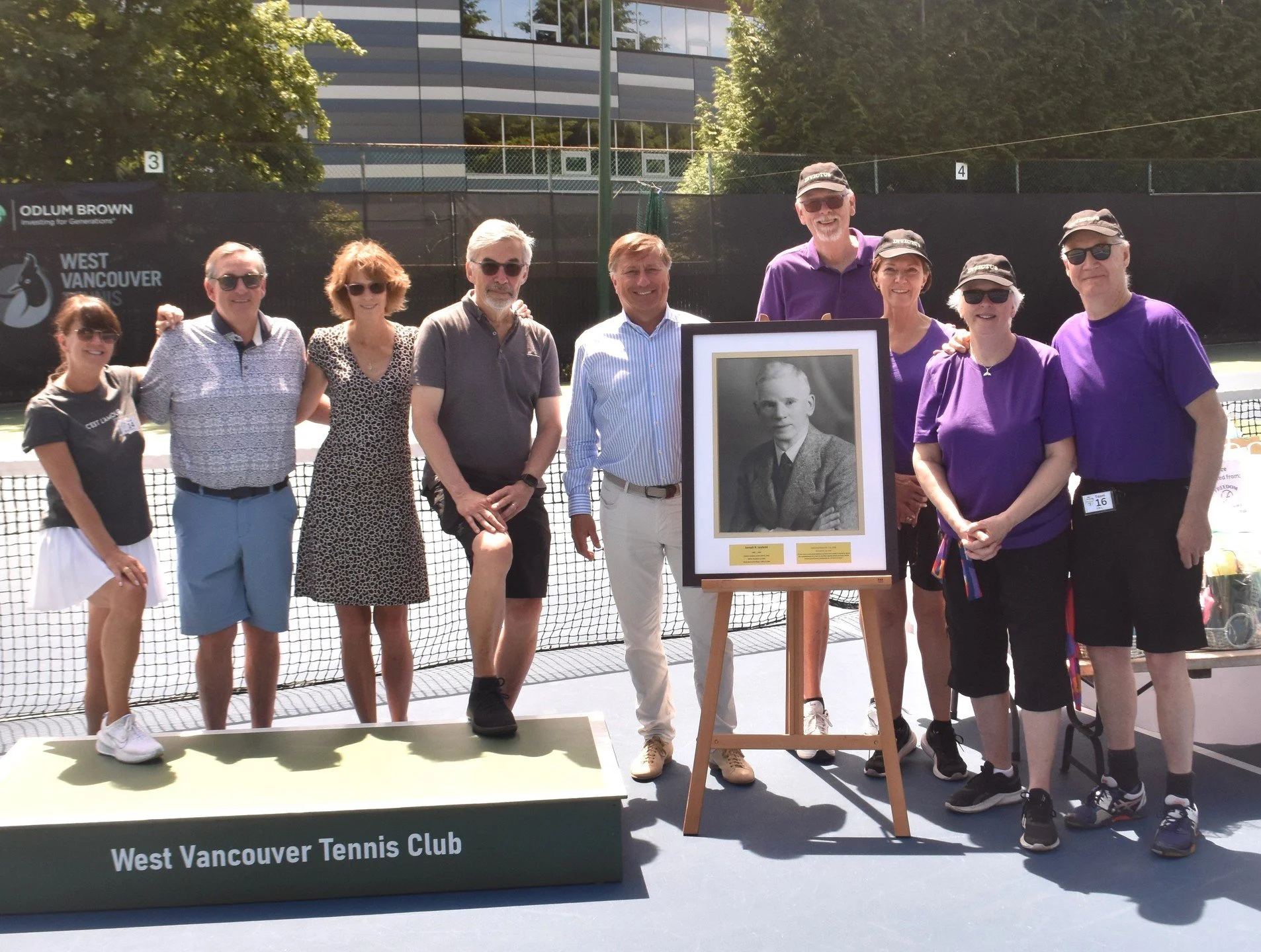 A group of ten people standing on a tennis court at the West Vancouver Tennis Club, posing for a photo around a framed black-and-white portrait of a young boy. Some are wearing sunglasses and hats, dressed in casual sports attire, with the background showing trees and a modern building.
