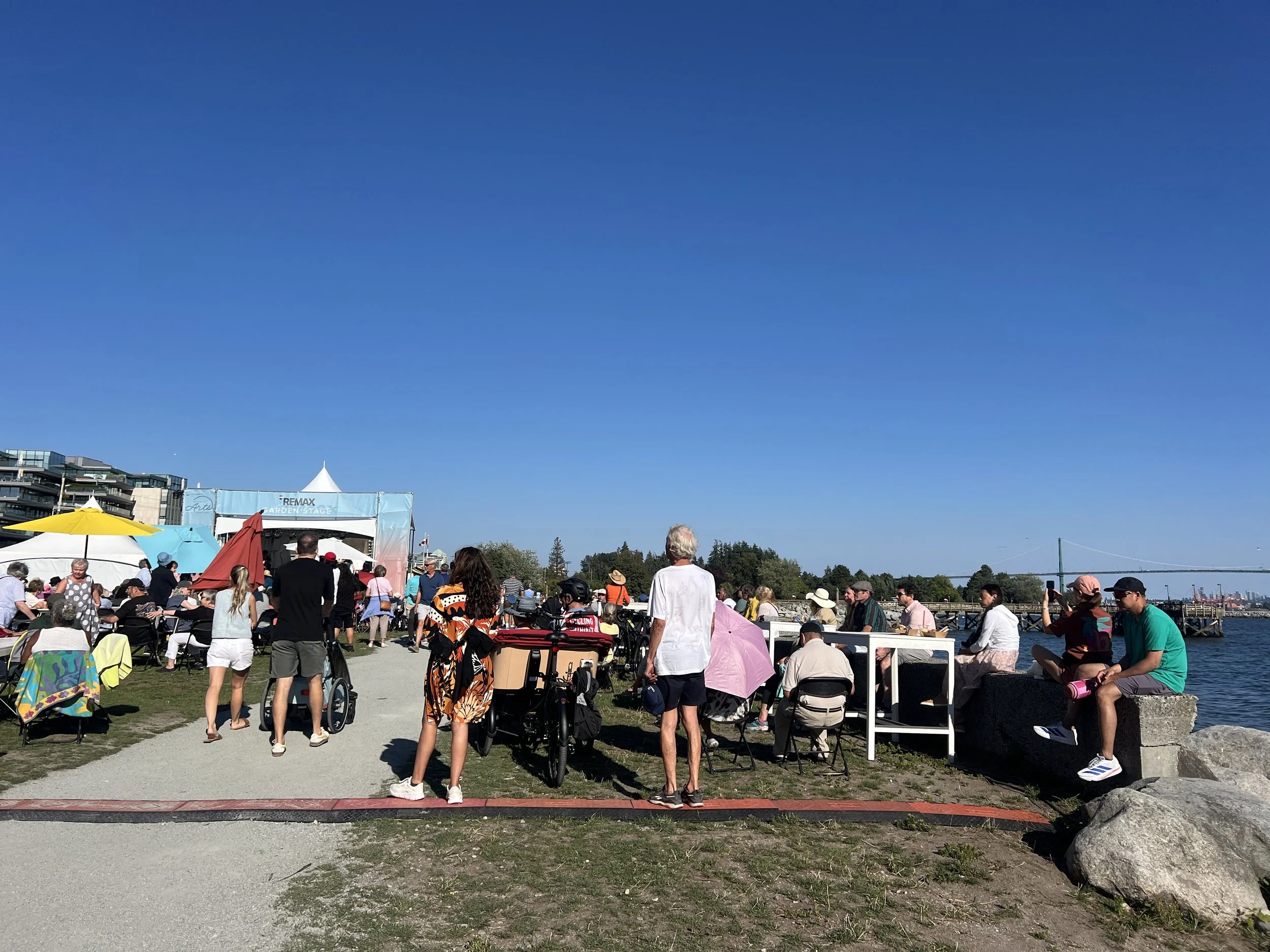 People gather outdoors near water, sitting on benches and standing, some with umbrellas, at a sunny event on a grassy area with tents and tall buildings in the background.