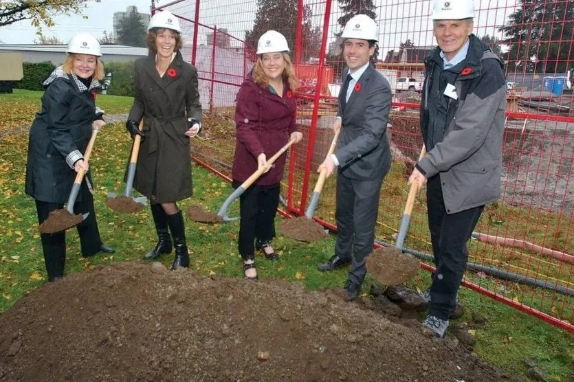 Five people, four women and one man, wearing hard hats and holding shovels, standing in front of a red construction fence, planting or digging in a dirt pile at a construction site