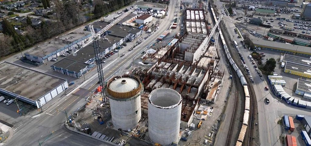 An aerial view of a construction site with two large cylindrical cooling towers, adjacent to railway tracks with train cars, roads with vehicles, and industrial buildings.