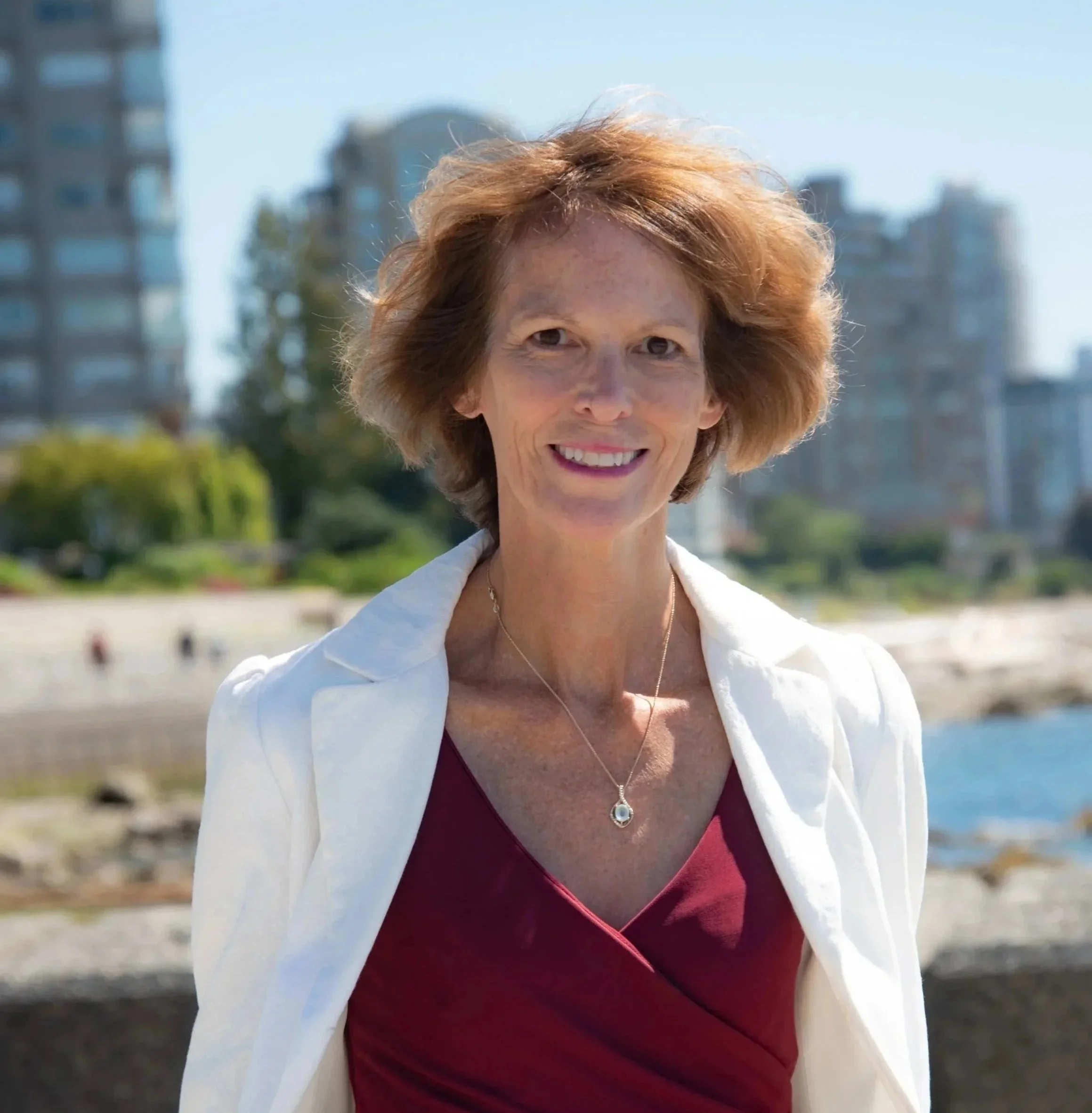 A woman with short red hair smiling outdoors with buildings in the background.