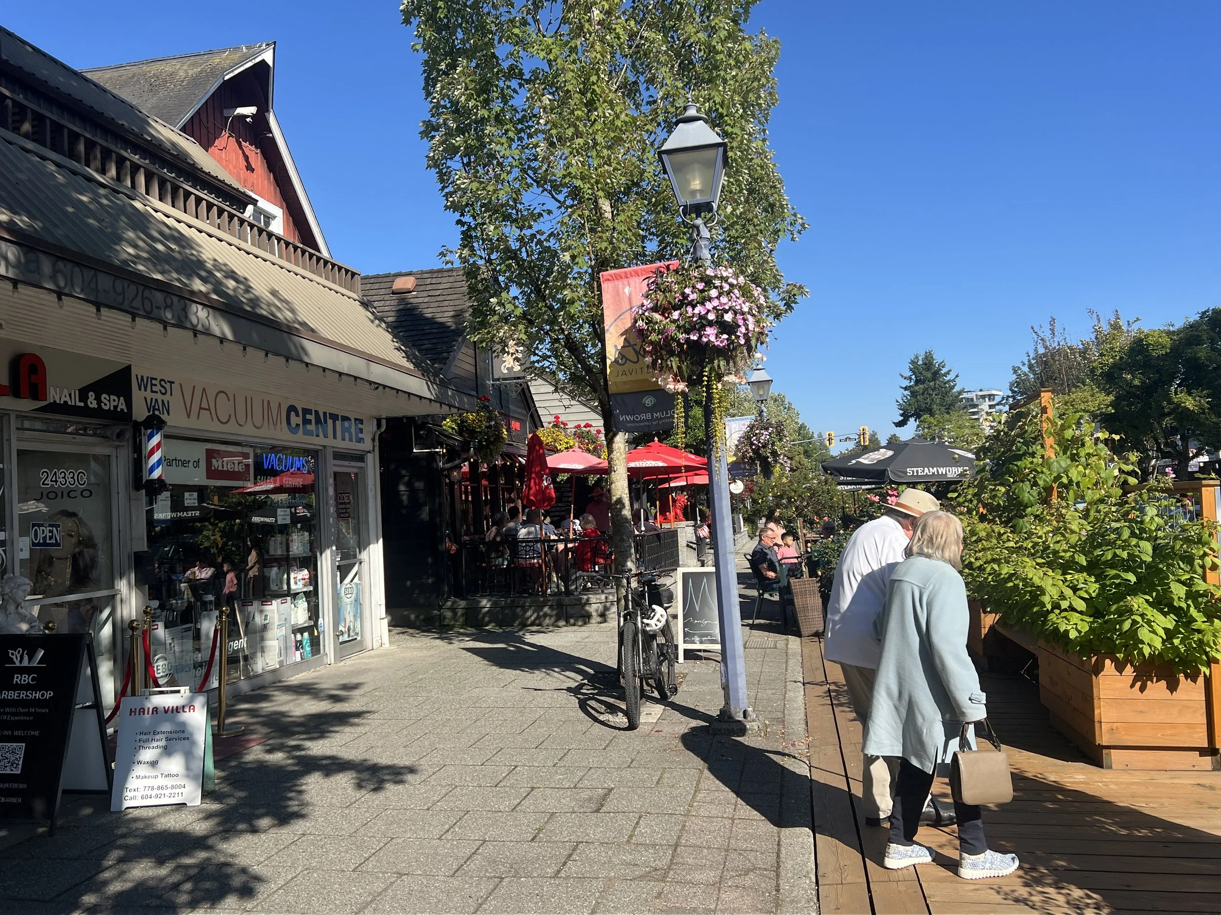 A lively downtown street scene on a bright, sunny day with storefronts, outdoor seating with red umbrellas, pedestrians walking, hanging flower baskets, trees, and blue sky.