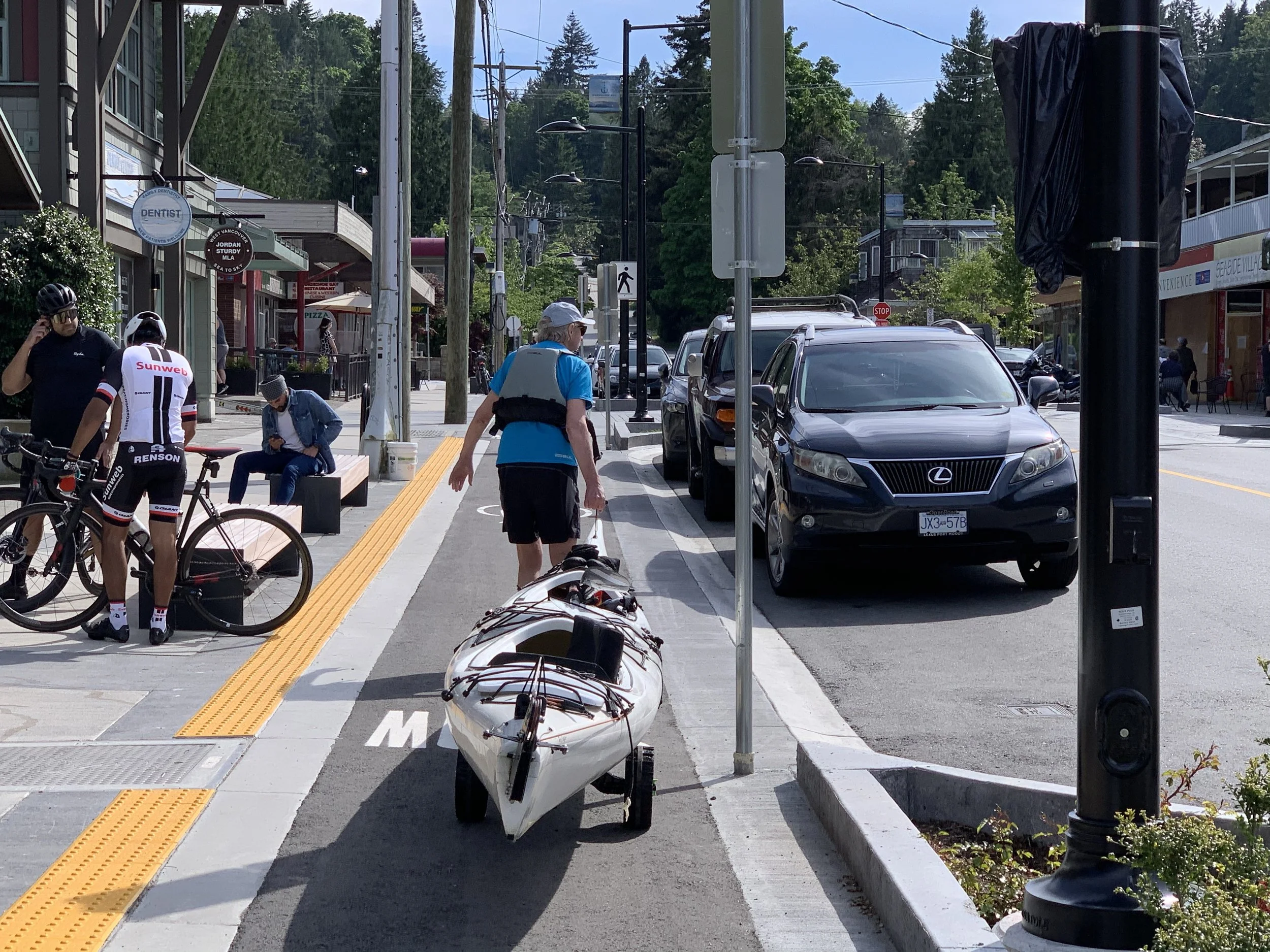 A city sidewalk with a kayak parked on the curb, people walking and standing, some wearing cycling gear and helmets, parked cars, buildings, and trees in the background.