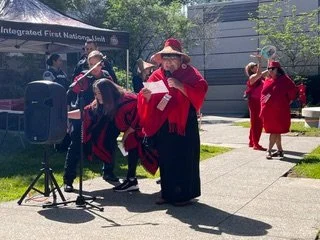 Group of people, including a woman with a red scarf and hat, reading from a paper at an outdoor event near a Fruit National Historic site, with others socializing nearby.