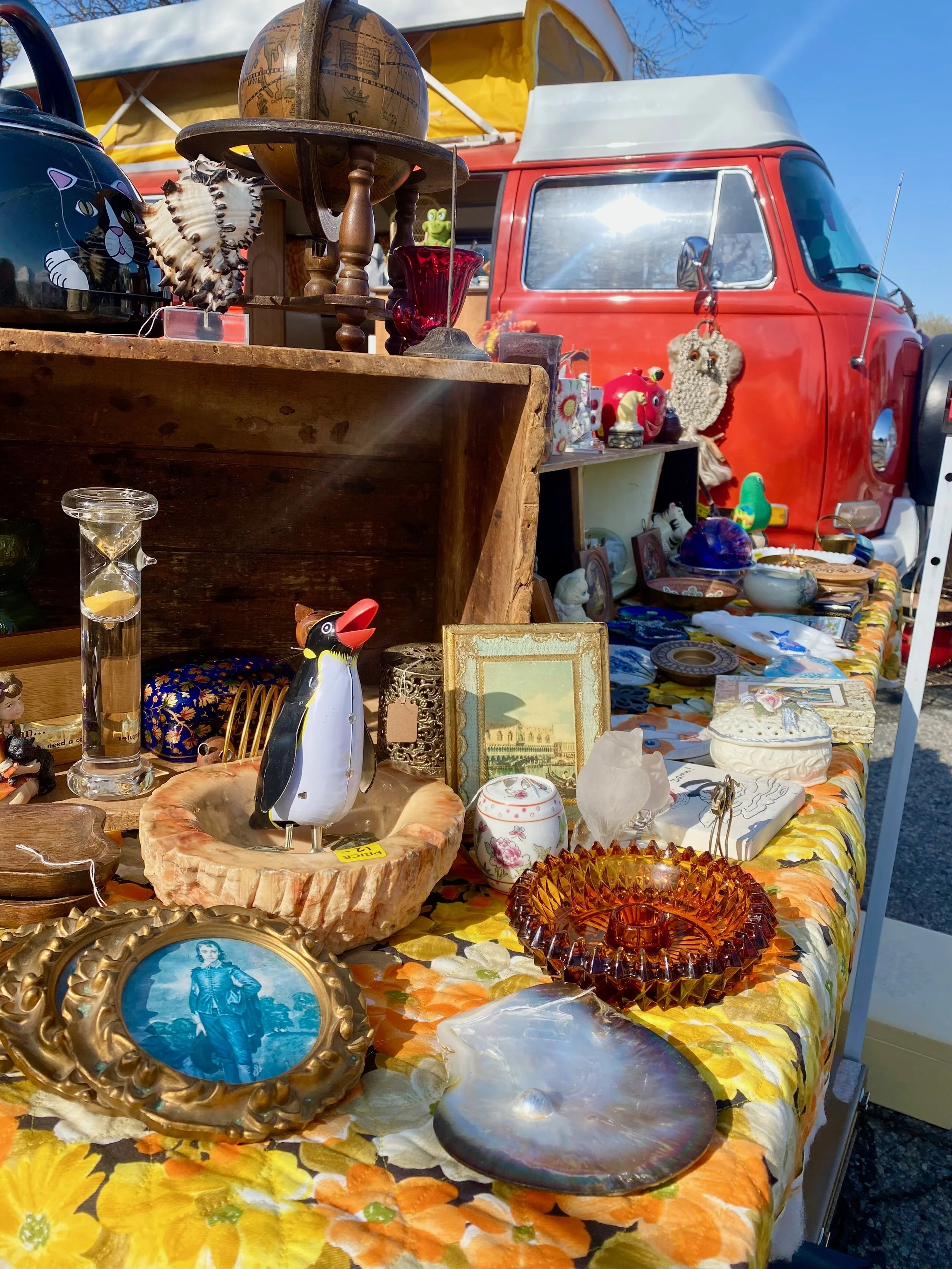 Table at a flea market with various vintage and decorative items, including picture frames, glassware, figurines, and ceramics, set against a background of a red vintage van and a clear blue sky.