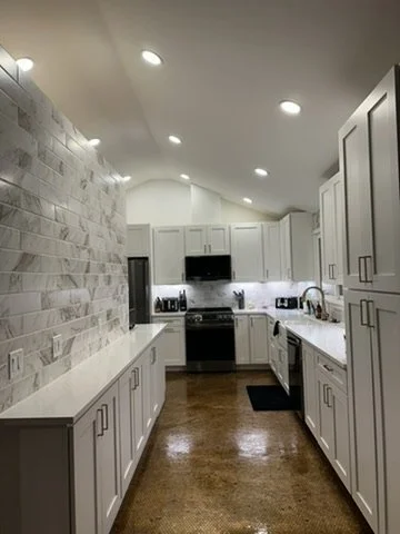 Modern kitchen with white cabinets, stainless steel appliances, wooden floors, and a stone accent wall with recessed lighting.