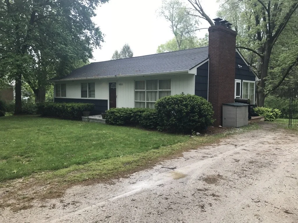 A single-story house with a dark gable roof, white and navy exterior, large front window, front door, brick chimney, surrounded by green bushes and trees, with a gravel driveway.