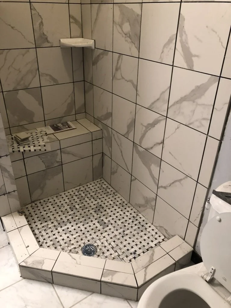 A tiled shower area with marble-patterned tiles, a built-in seat with a stack of pamphlets or magazines, a small shelf, and a drain, located next to a toilet.