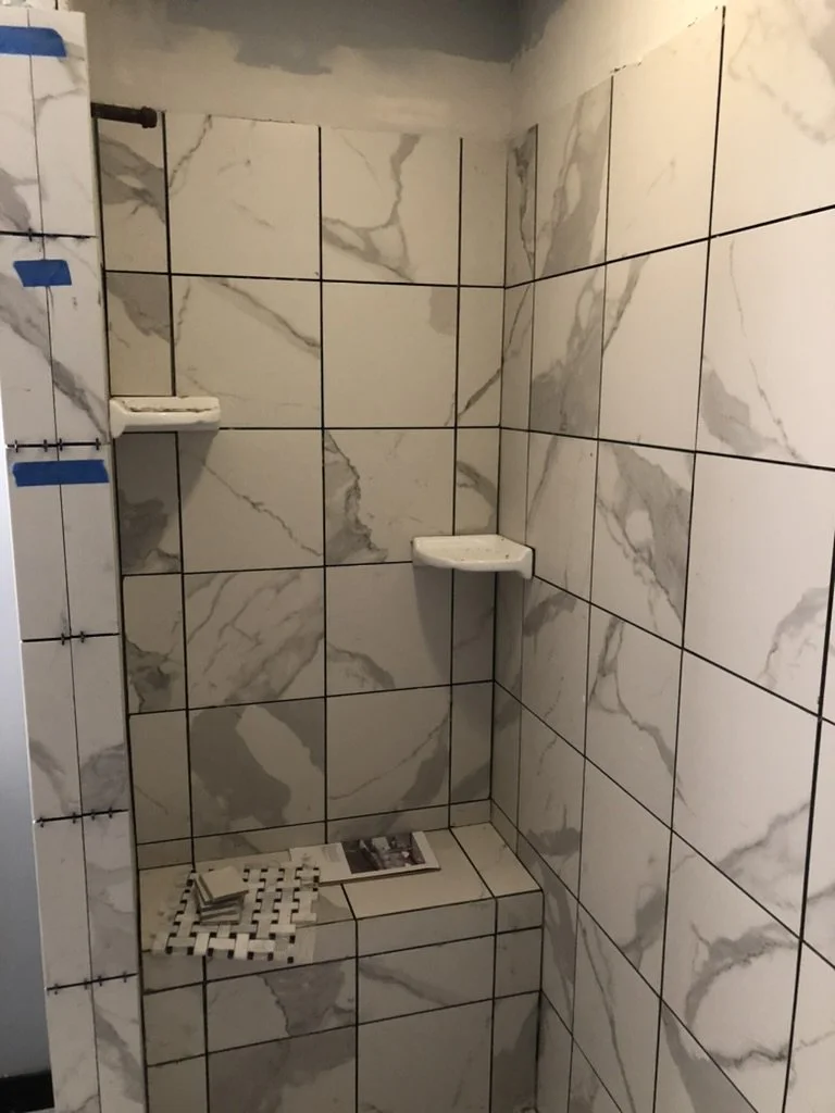A shower area with marble-patterned tiles, two corner shelves, and a built-in tiled seat, with some tiles, a newspaper, and cleaning supplies on the seat.