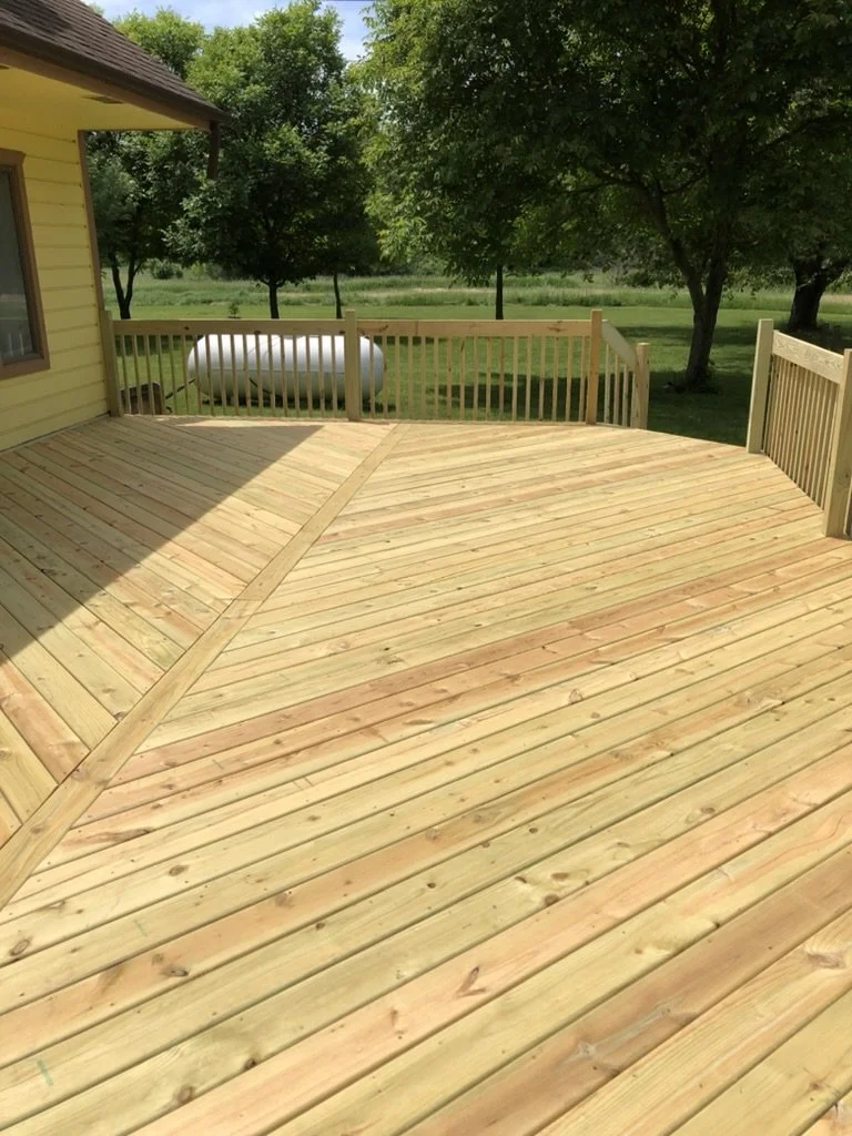 A newly built wooden deck attached to a yellow house in a backyard, surrounded by green trees and a grassy lawn, with a propane tank in the background.